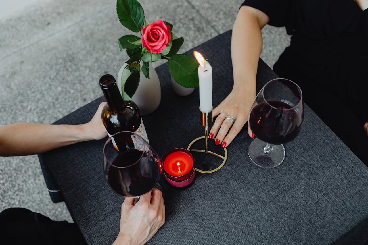 Two people sitting at a table with a red rose in a vase, a lit candle, and glasses of red wine. One person holds a bottle, while the other has a hand near a candle. Theres a romantic ambiance.