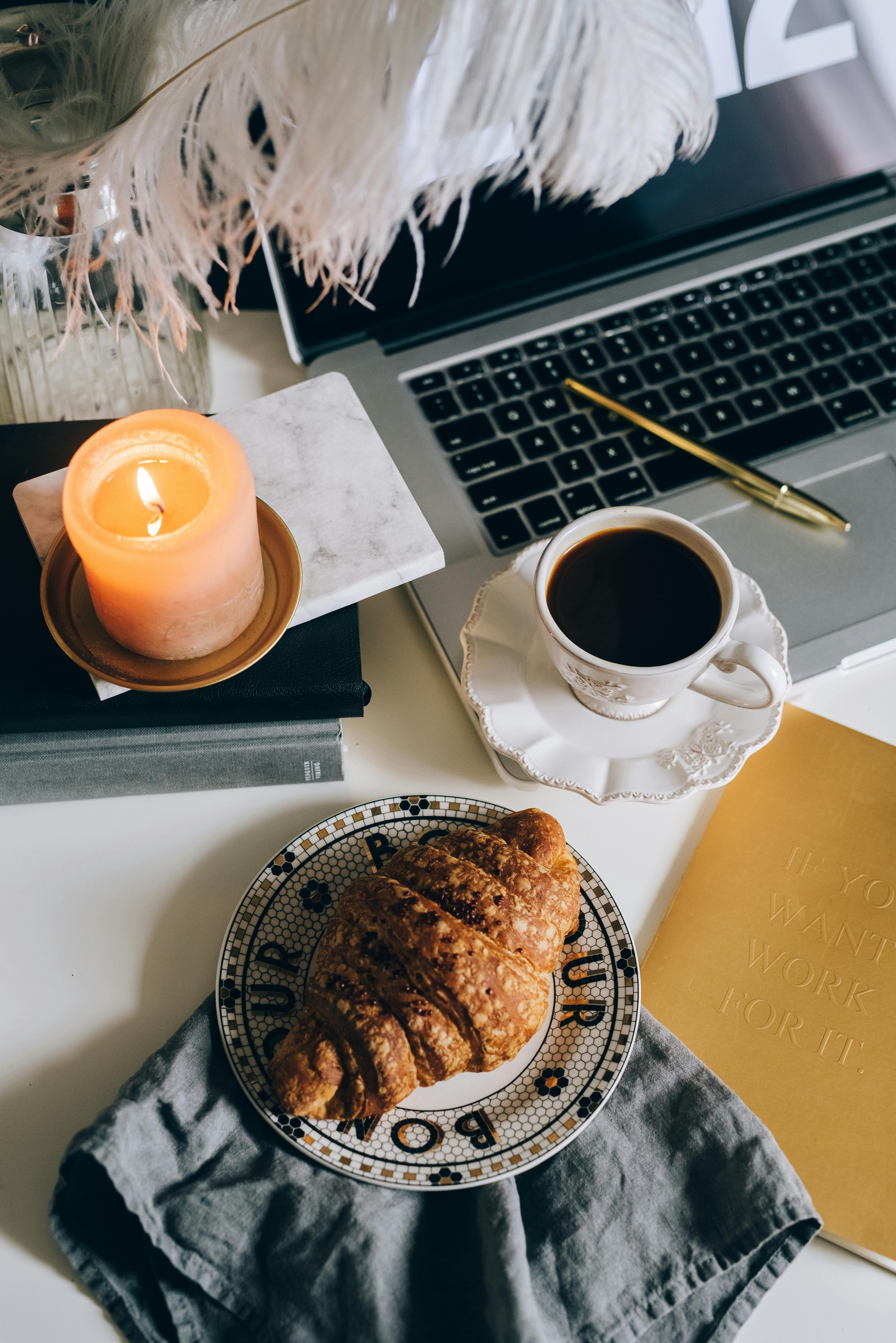 A croissant on a patterned plate, a cup of coffee, a lit candle, an open laptop, and a yellow notebook on a desk with a gray napkin and decorative feathers overhead.