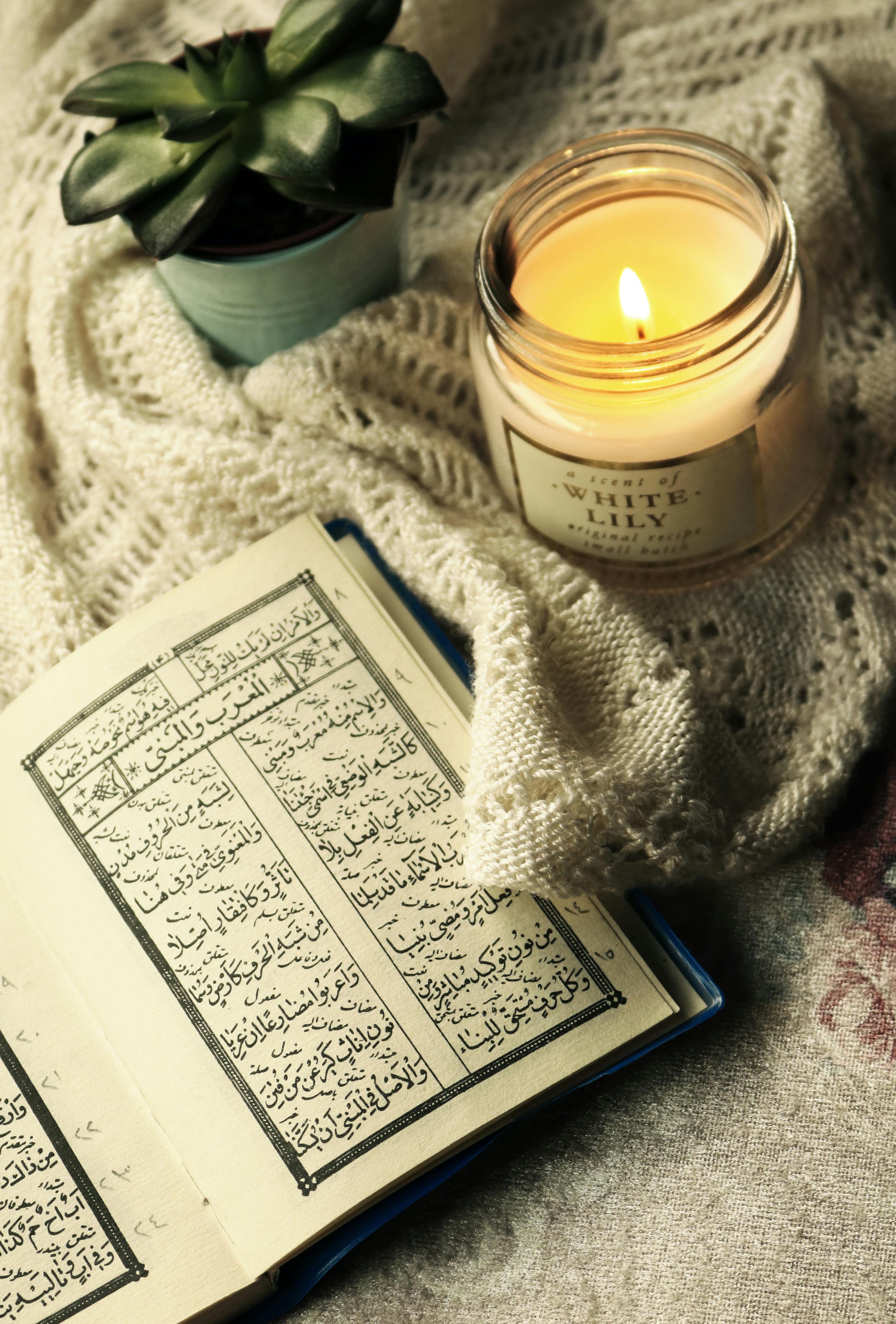 An open book with Arabic text lies on a knitted cloth next to a lit white candle labeled “White Lily” and a small potted succulent plant.