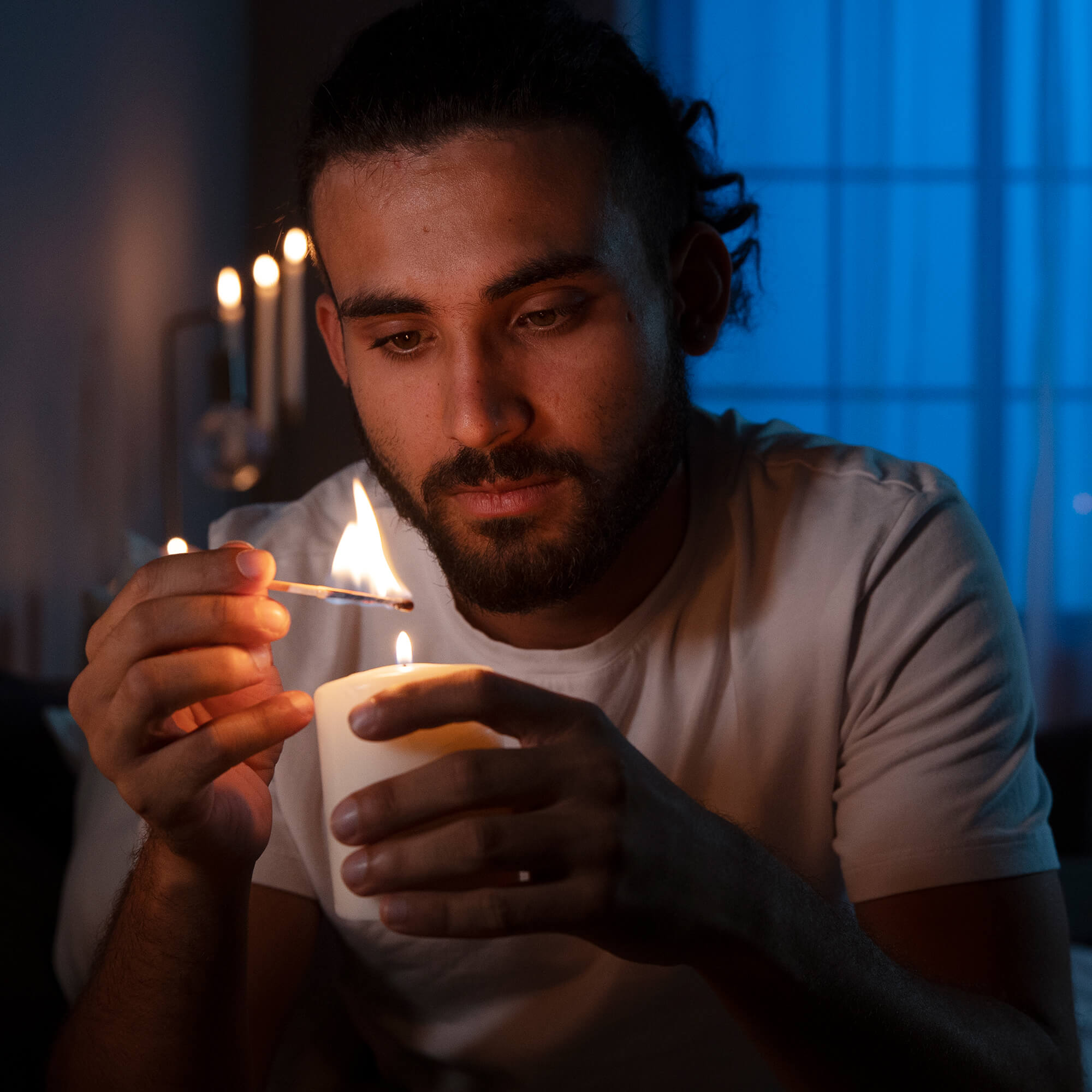 A person with a beard and tied-back hair lights a white candle with a match while sitting indoors. The room has a soft blue and warm yellow glow from candlelight.