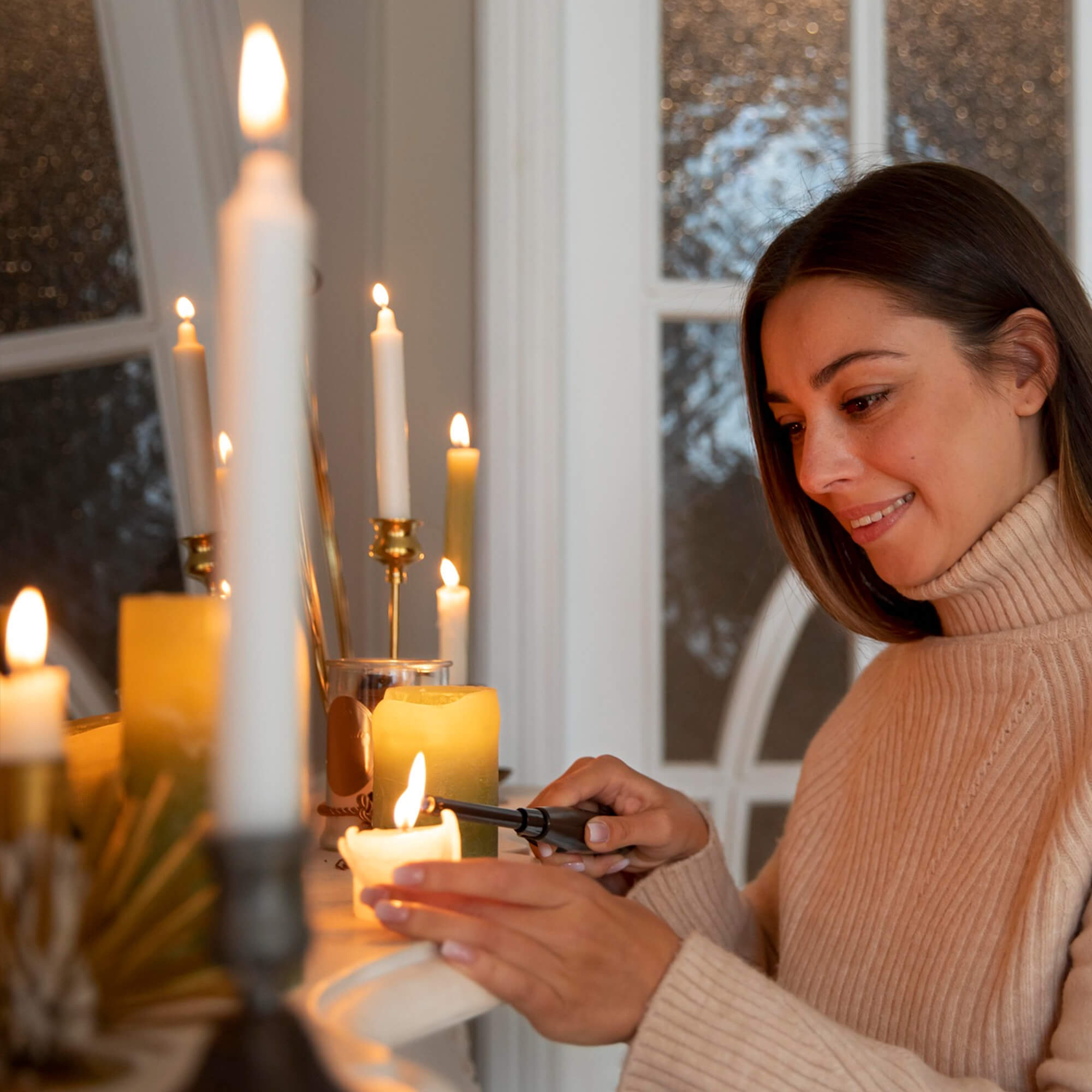 A woman in a cozy sweater lights candles on a mantelpiece with a lighter. The room has a warm and serene ambiance, with several lit candles of various sizes creating a soothing atmosphere.