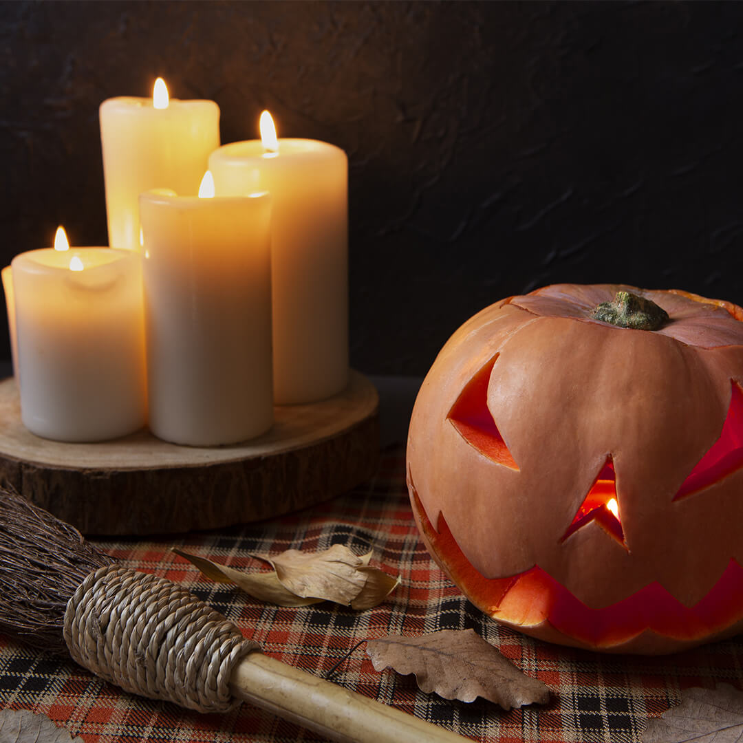 A carved pumpkin with a spooky face glows beside a group of lit candles, resting on a wooden base. A straw broom and fallen leaves are nearby, all set against a dark background.