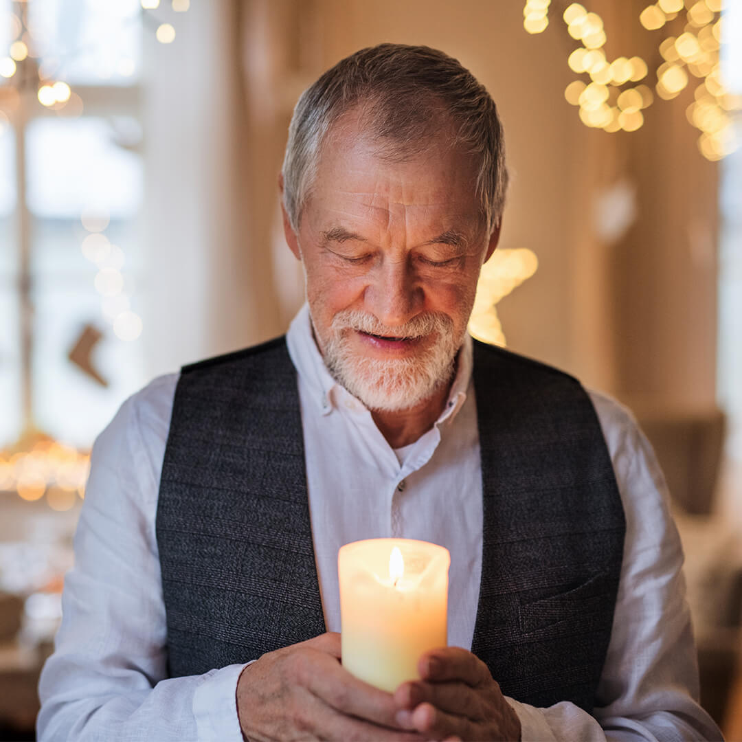 An elderly man holds a lit candle with both hands, smiling gently. The background is softly lit with blurred fairy lights, creating a warm and cozy atmosphere.