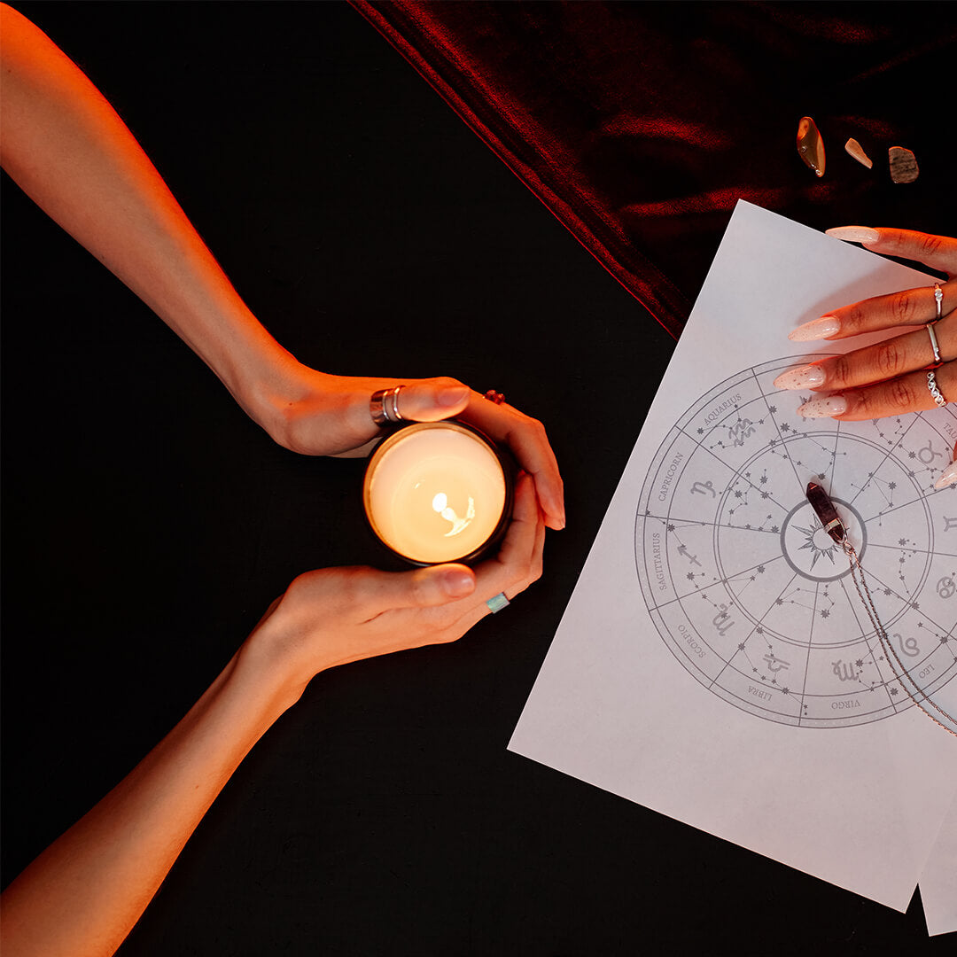 Two hands with rings are visible; one holds a lit candle, the other points at a zodiac chart on a table. Dim lighting creates a mystical atmosphere with crystals nearby.