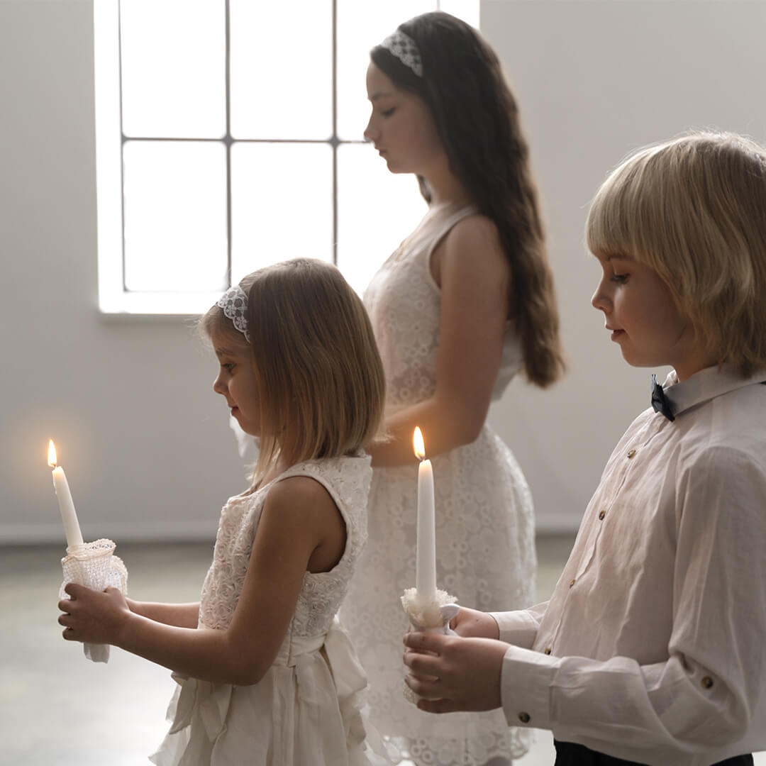 Three children in white clothing hold lit candles in a calm, softly lit room. They stand in a line, gazing at the flames, with a large window in the background providing natural light.