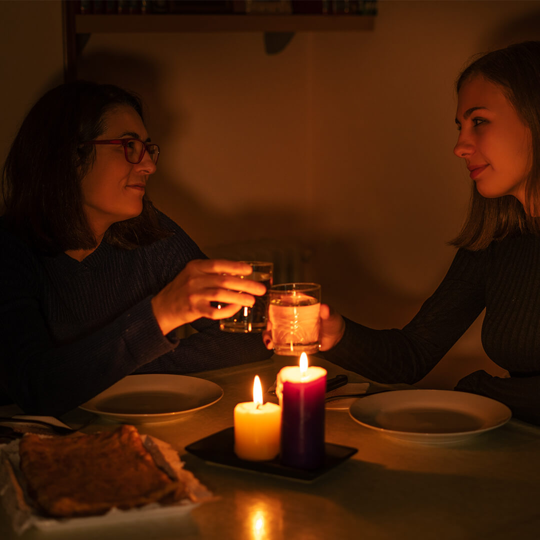 Two people sitting at a dimly lit table, raising glasses in a toast. Three candles illuminate the scene. A dish of food is placed on the table, creating a cozy atmosphere.