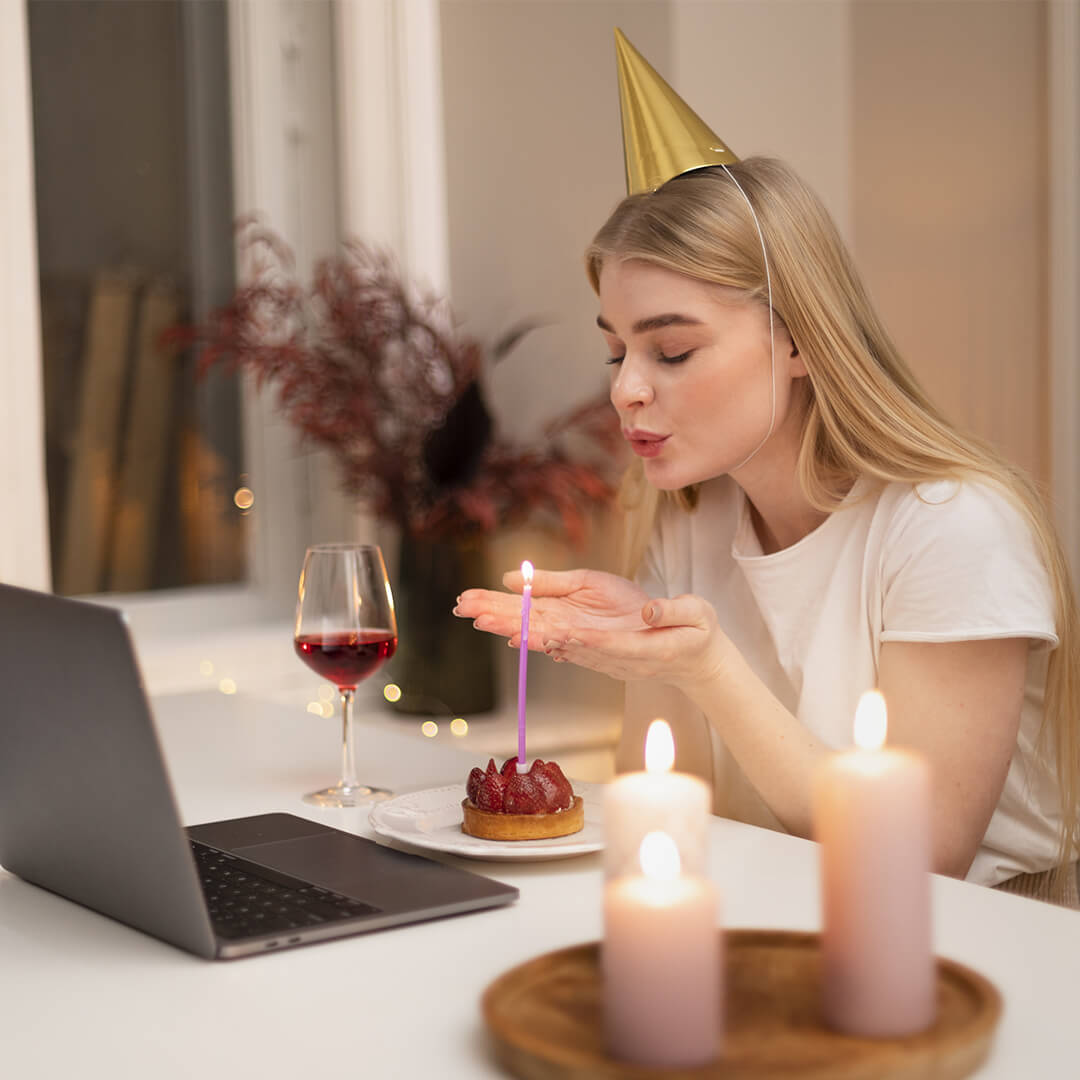 A woman wearing a party hat blows out a candle on a dessert. She is sitting at a table with a laptop, a glass of red wine, and lit candles. The setting appears to be a cozy indoor celebration.