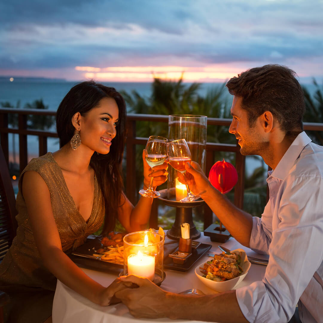 A couple toasts with wine glasses at a candlelit dinner on a balcony overlooking the ocean at sunset, creating a romantic ambiance.