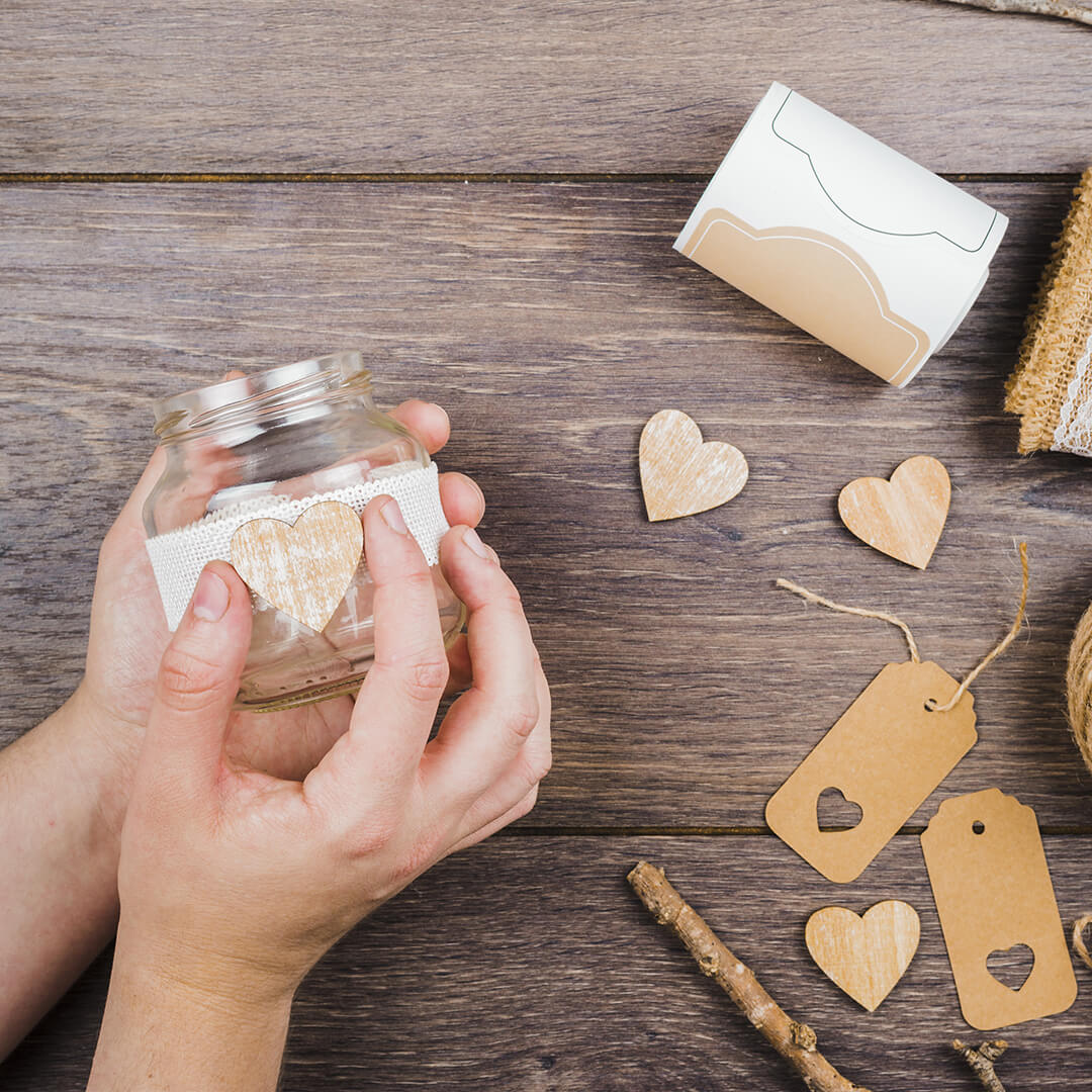 Hands decorating a glass jar with a lace band and heart-shaped sticker on a wooden table. Surrounding items include heart cutouts, tags, and a roll of decorative tape.