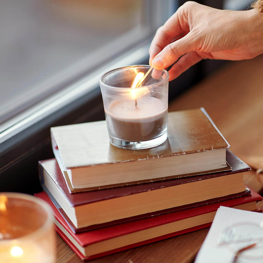 A hand lighting a candle placed on a stack of books near a window. Another candle is softly glowing in the background, creating a cozy atmosphere.