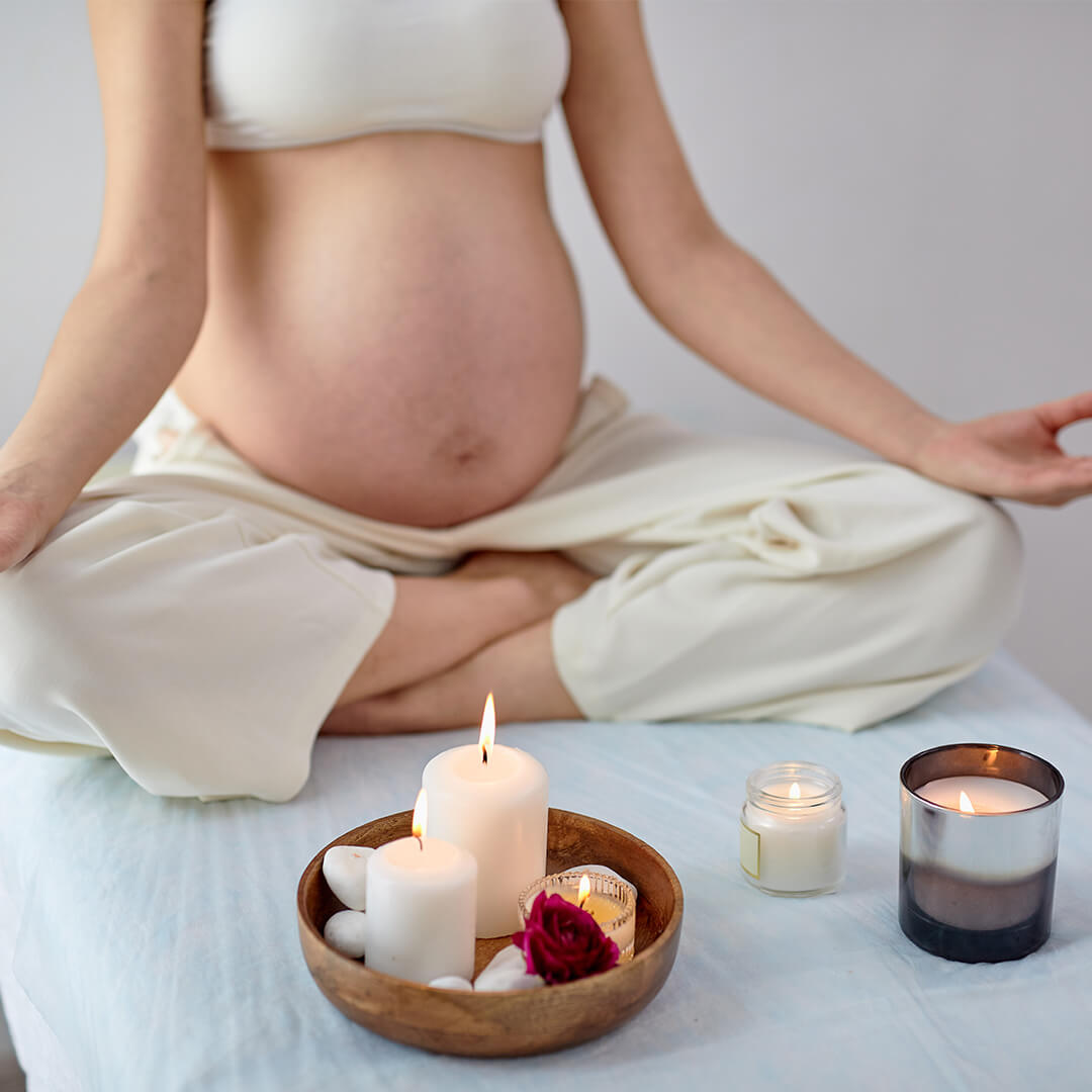 A pregnant person sits cross-legged in meditation, wearing a white top and pants. Nearby are lit candles in jars and a wooden bowl with stones and a rose, creating a calm setting.