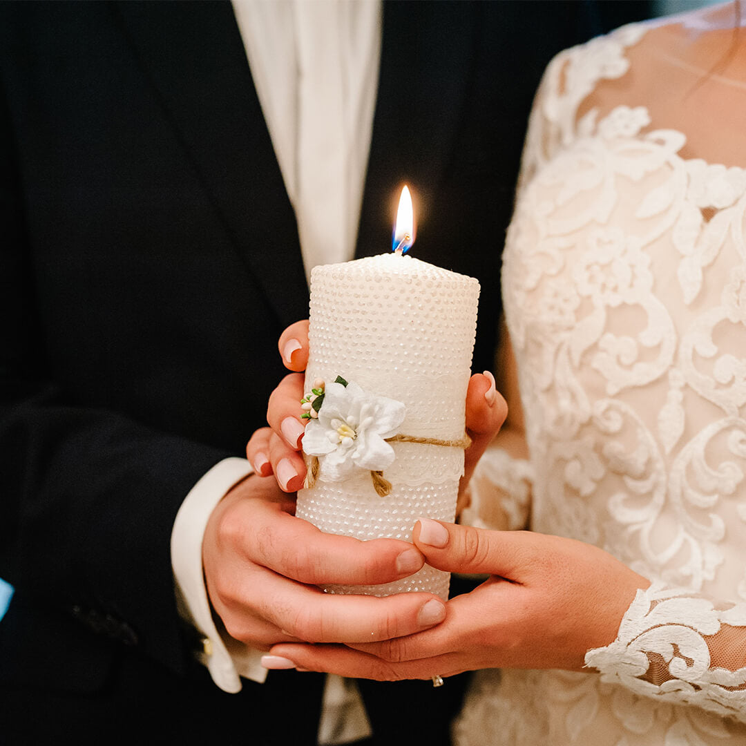 A couple in formal attire holds a lit candle decorated with flowers, symbolizing unity. The woman wears a lace wedding dress, and the man wears a dark suit.