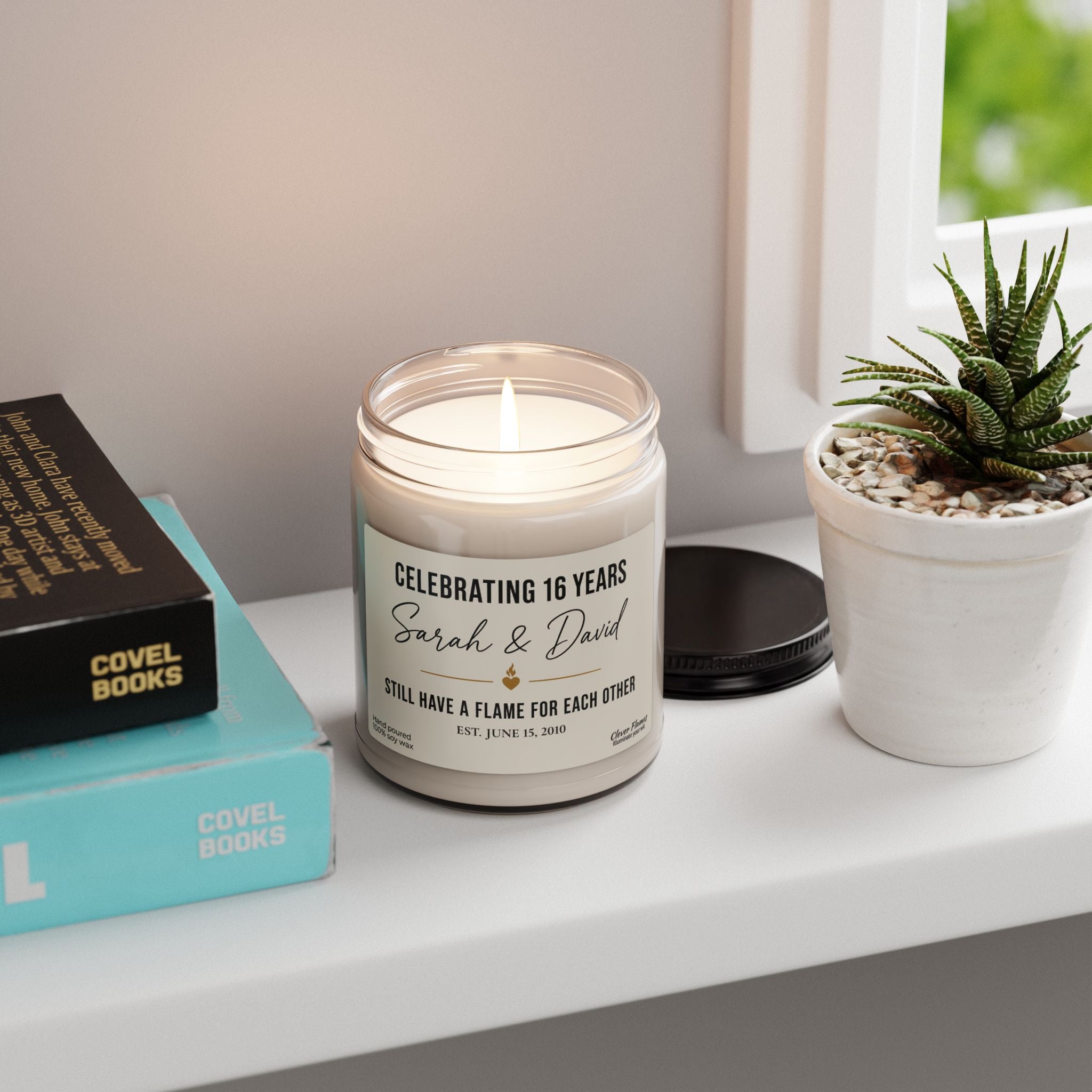 The Personalized 16th Anniversary Candle by Printify—a custom wax anniversary gift for couples celebrating 16 years married—rests on a white shelf next to stacked books, a sleek black lid, and a small potted succulent by the window. Featuring a unique custom label in a classic glass jar, this thoughtful gift for husband or wife adds warmth and charm to any space.