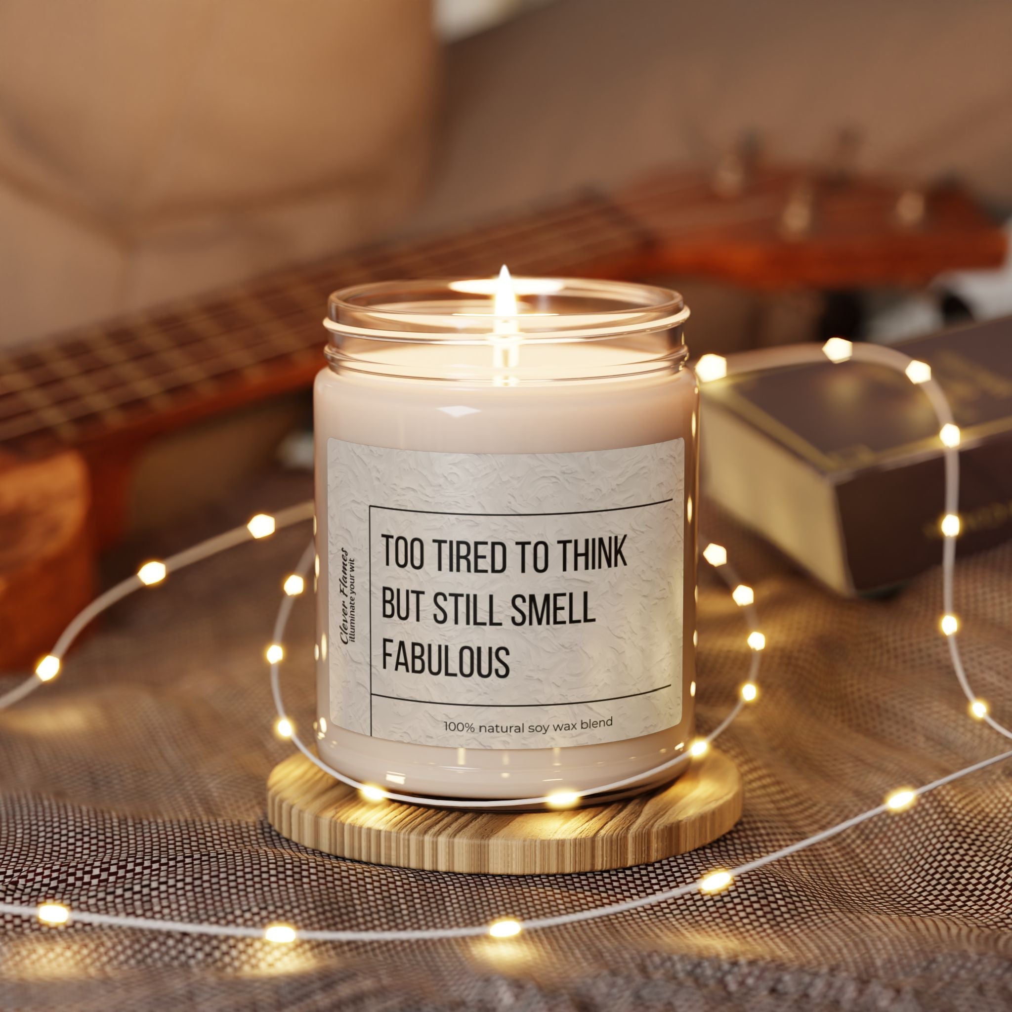 A lit soy candle in a glass jar, labeled as the Too Tired to Think Candle - Smells Like Fabulousness from Printify, sits on a wooden coaster. Surrounded by glowing string lights and a small book, an unfocused ukulele rests in the background, adding to the eco-friendly ambiance.