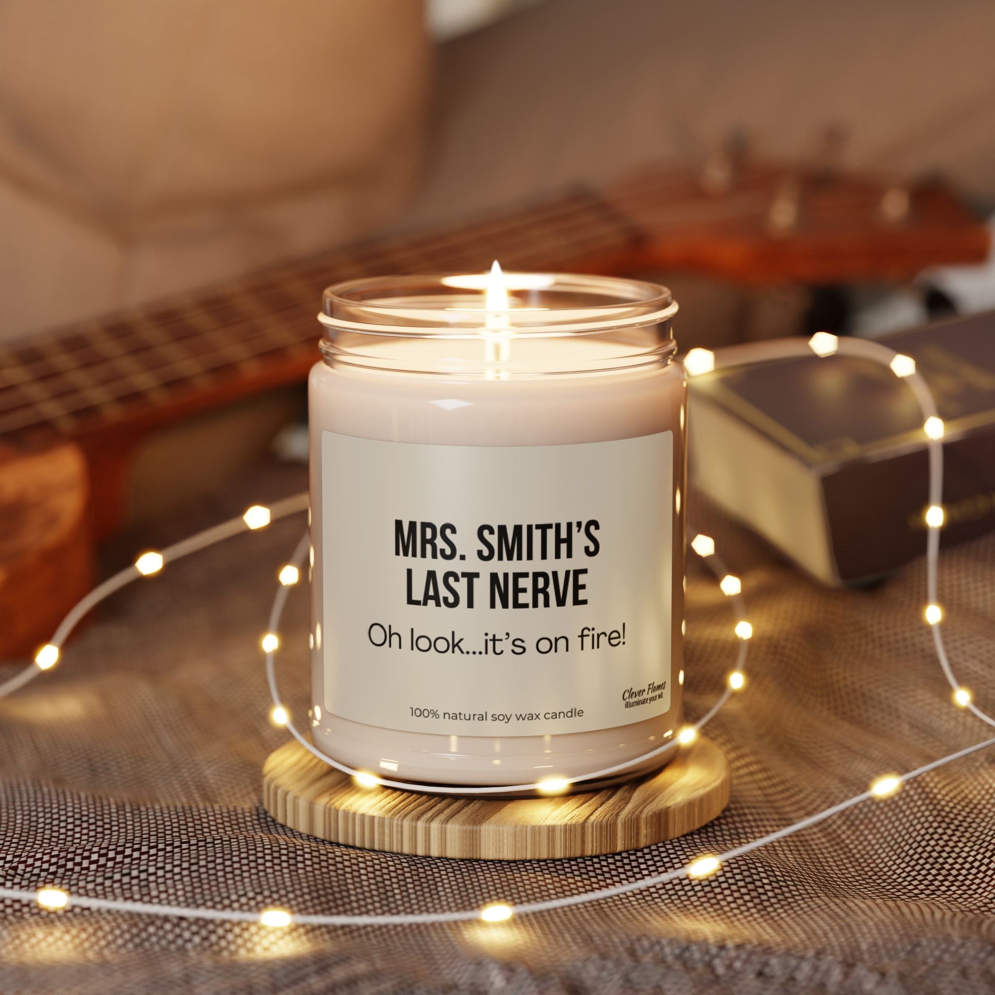 A glowing soy candle from Printify, personalized with "Teacher Appreciation Last Nerve" and the humorous phrase "Oh look...it's on fire!" sits elegantly on a wooden base. Encircled by twinkling fairy lights, it serves as an ideal gift for teachers, set against a softly blurred backdrop featuring a guitar and gentle fabric.