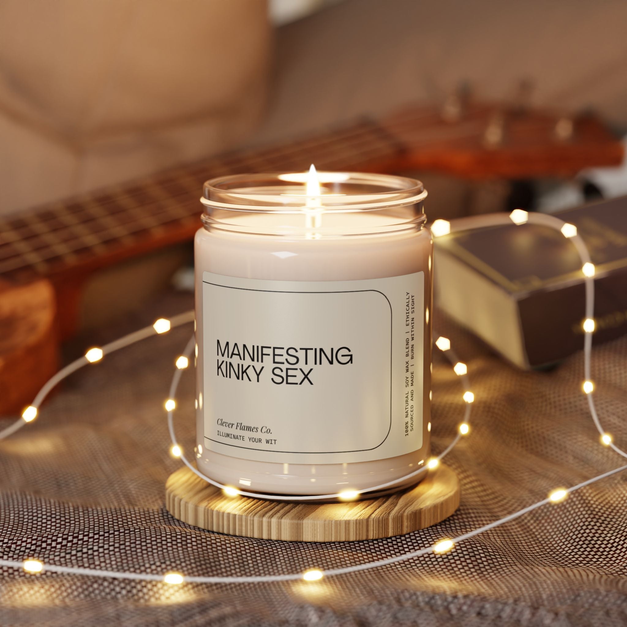 A soy candle titled "Manifesting Kinky Sex" by Printify is perched on a wooden coaster. It is enveloped by a strand of small glowing lights atop a textured surface, while a blurred guitar and book in the background add to the cozy atmosphere.