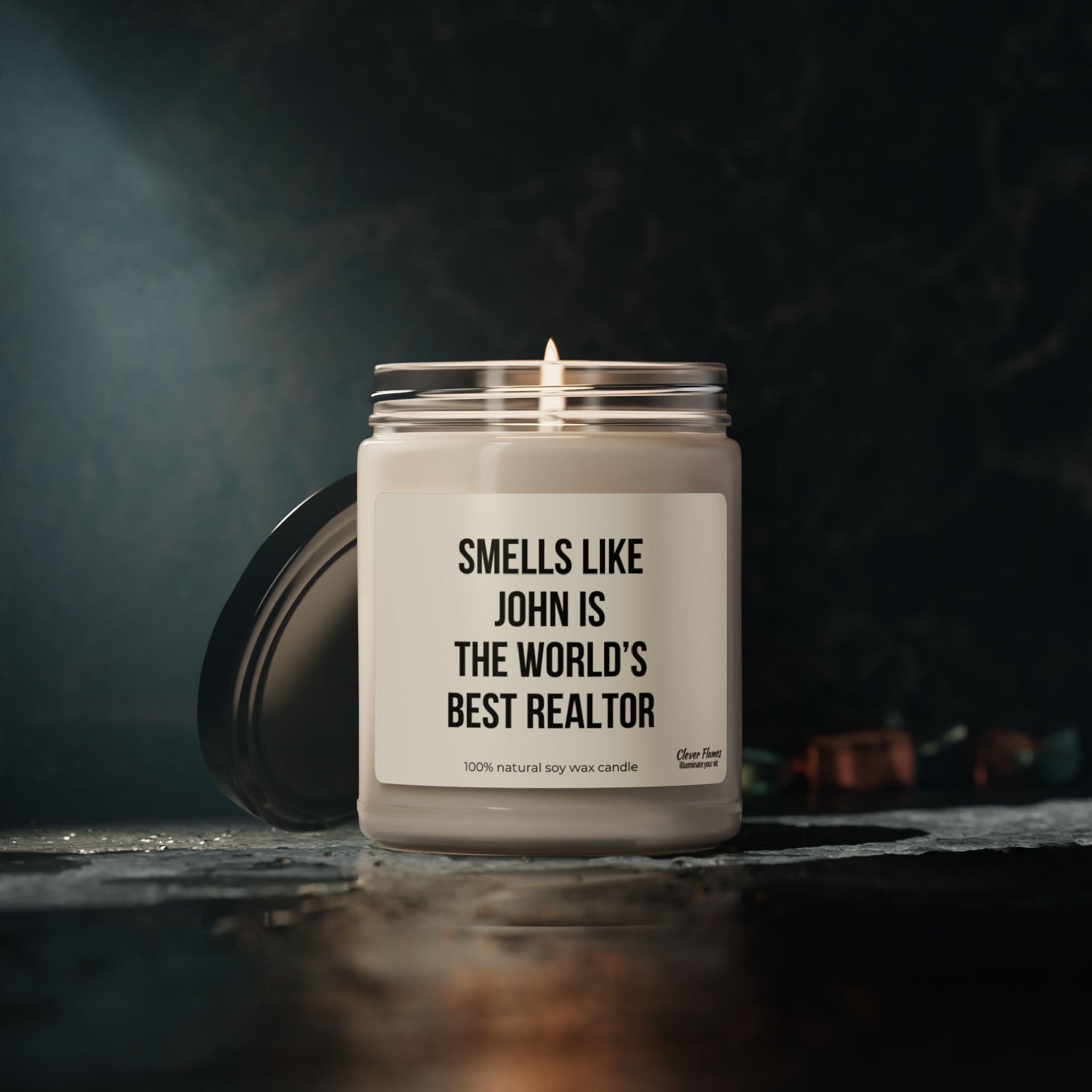 A customized Printify candle jar with a black lid sits on a dark surface, its label declaring, "Smells Like World's Best Realtor." Crafted from natural soy wax, it's an ideal gift for real estate professionals. The dimly lit backdrop accentuates its cozy glow.