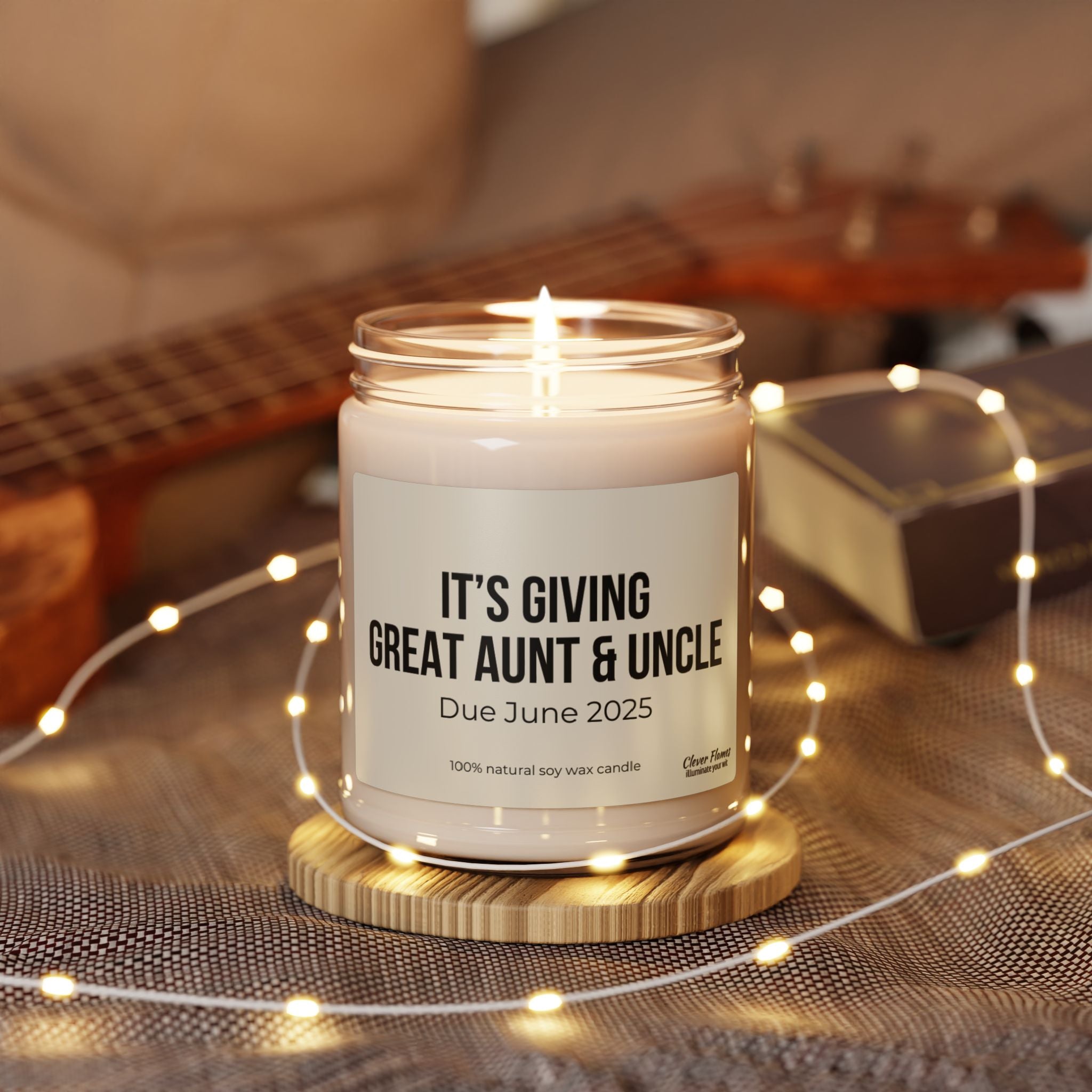 A lit candle from Printify, named "Great Aunt and Uncle Pregnancy Announcement Candle," inscribed with "Due June 2025," sits on a wooden coaster. Surrounded by fairy lights, it brings charm to the blurred background featuring a ukulele and a gift box—an ideal personalized gift choice for a baby reveal or pregnancy announcement.