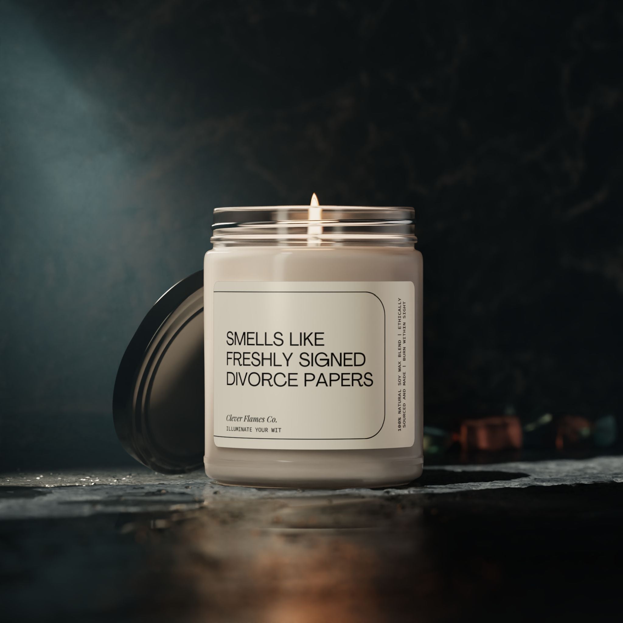 A lit candle in a jar labeled "Smells Like Freshly Signed Divorce Papers" from Printify sits on a dark surface, making it the perfect humorous divorce gift. Crafted from natural soy wax, this scented candle emits a warm, soft glow and is an ideal breakup or funny gift for women.