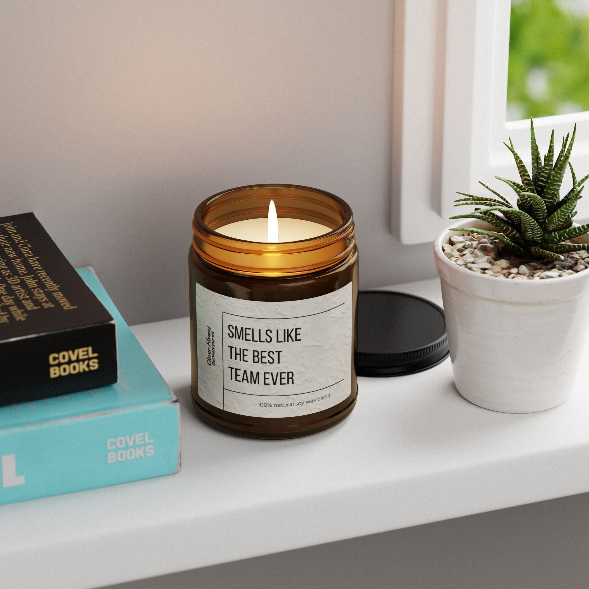 A lit brown glass candle from Printify, labeled as a personalized appreciation gift for the best team ever and crafted from eco-friendly scented soy wax, sits on a white surface beside a small potted plant and stacked books. In the background, you can see part of a white window frame. The black lid is removed and placed behind the candle.