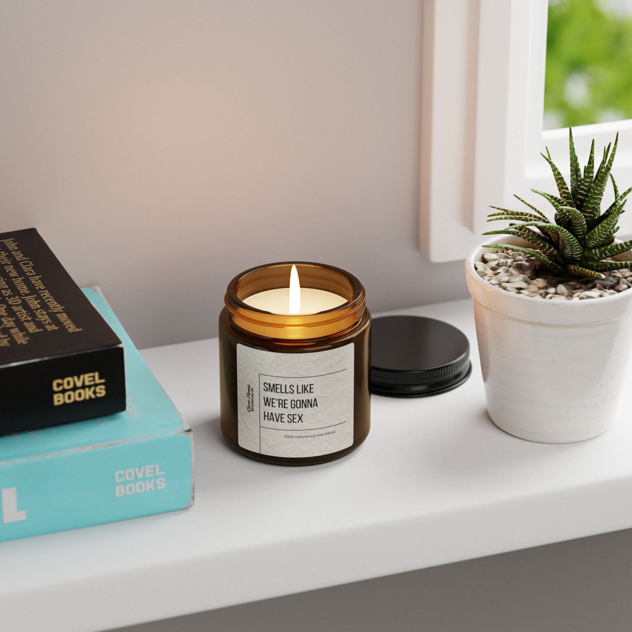 A Printify Smells Like Were Gonna Have Sex candle in a brown reusable glass vessel sits on a windowsill next to a small potted succulent. Nearby, blue books are stacked together, creating an eco-friendly and cozy corner.