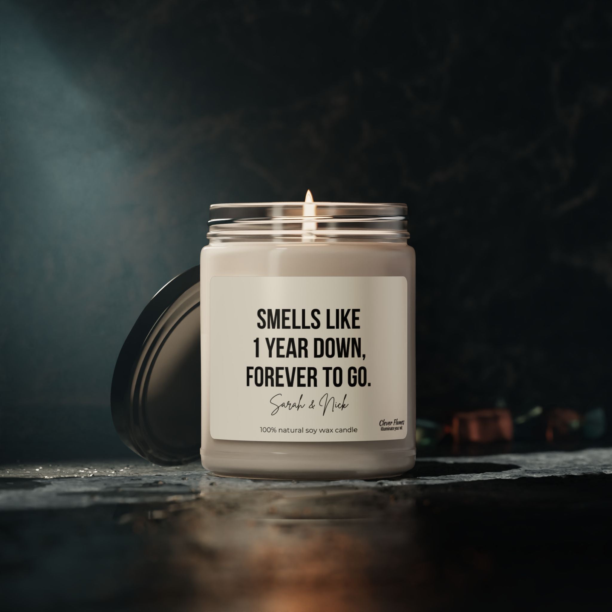 A personalized soy candle in a jar, branded by Wedding, rests on a dark surface, displaying the message "Smells like 1 year down, forever to go." The names Sarah & Nick below hint at its role as a custom anniversary candle celebrating their love. This makes for an ideal 1st anniversary gift, with the background gently blurred.