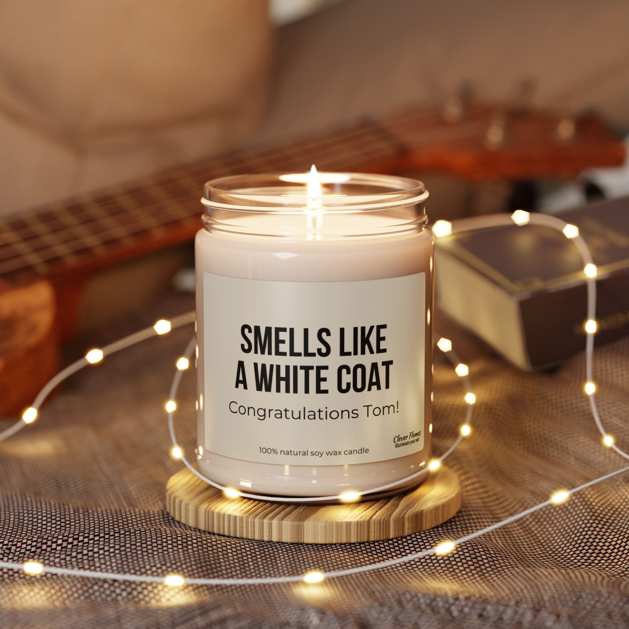 A personalized "Smells Like A White Coat" candle by Printify, designed for a doctor in training, flickers from its glass jar labeled "Congratulations Tom!" It's surrounded by gentle string lights, with a ukulele and book softly blurred in the background—an ideal gift for celebrating a medical student's white coat ceremony.