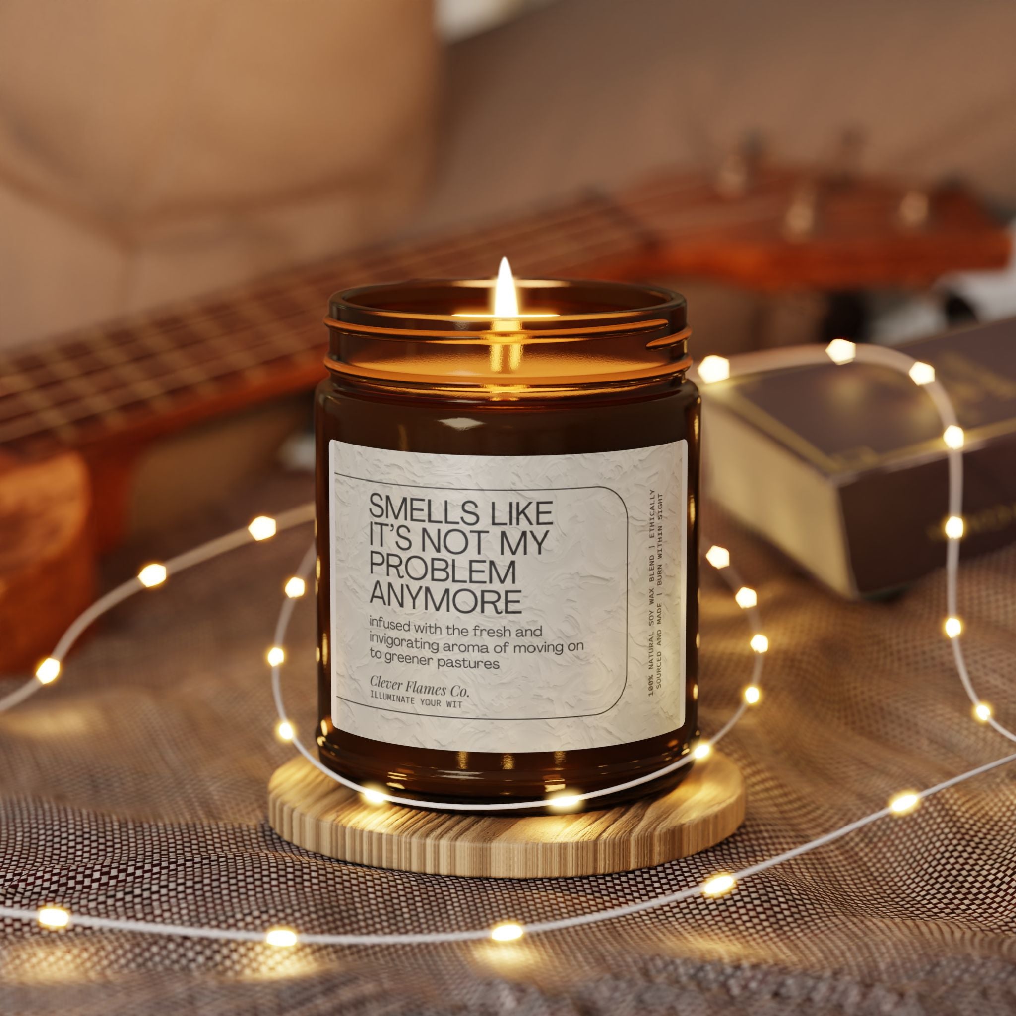 An illuminated, amber-colored "Funny Retirement Scented Soy Candle" by Printify, featuring a label saying "Not My Problem Anymore," rests on a wooden base. This eco-friendly soy candle is adorned with fairy lights on a textured surface, while a guitar and book appear softly blurred in the background.