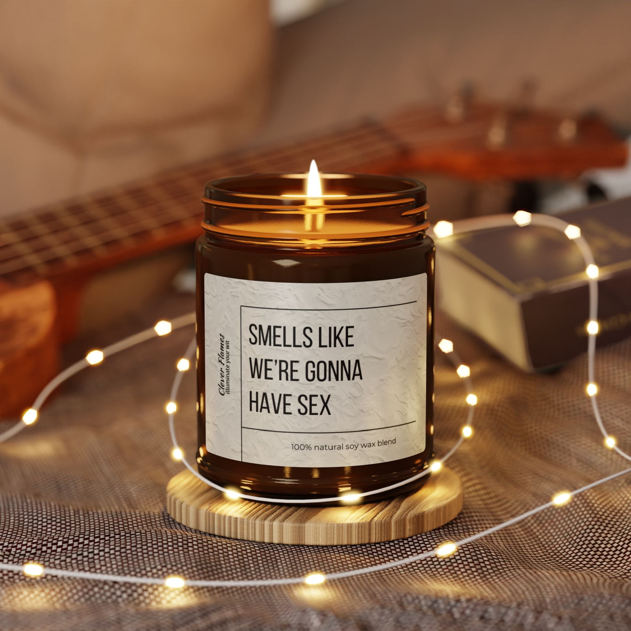 The Smells Like Were Gonna Have Sex candle by Printify, set in a reusable glass, rests on a wooden coaster with string lights, an out-of-focus ukulele, and book in the background, creating an eco-friendly and cozy vibe.