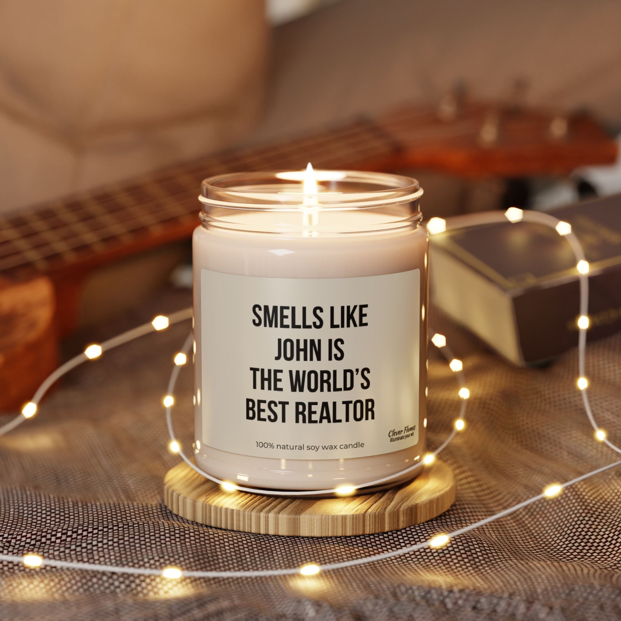 A Smells Like World's Best Realtor Candle by Printify, crafted from natural soy wax, sits on a knitted surface. Surrounded by fairy lights with a blurred ukulele in the background, it makes the perfect gift for real estate professionals.