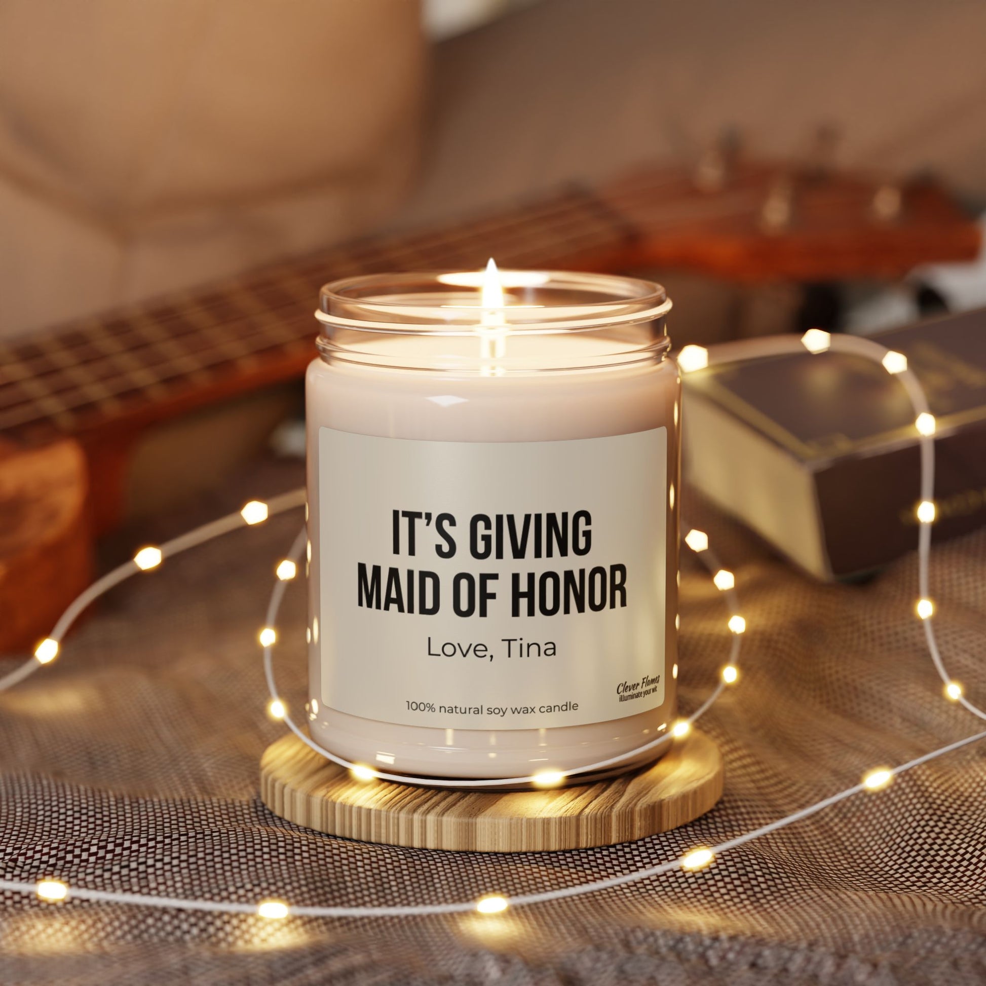 A personalized It's Giving Maid of Honor candle by Wedding, elegantly displayed on a wooden stand, makes an ideal gift for the maid of honor. Accompanied by fairy lights, a ukulele, and a book, it sets a cozy and warm ambiance.