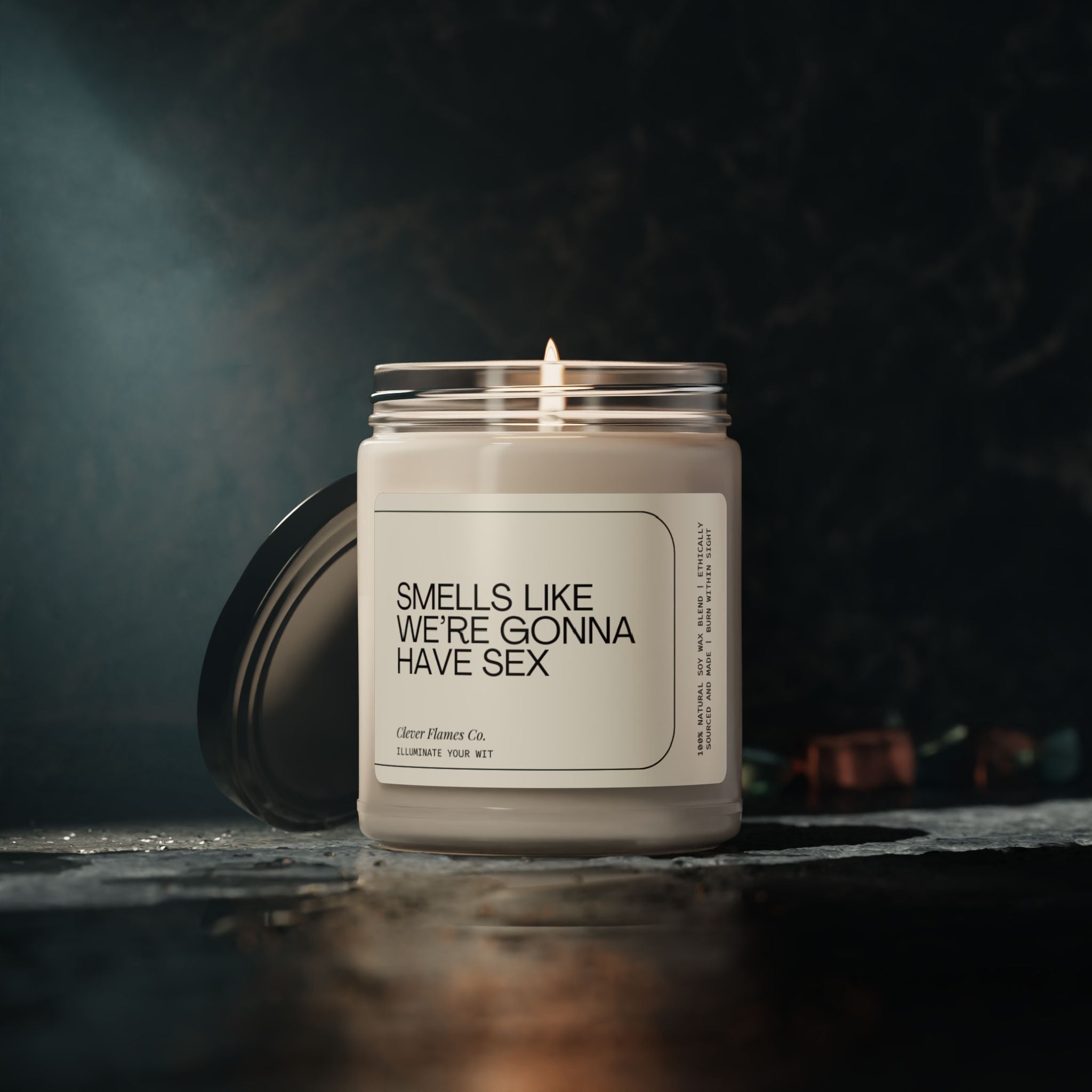 On a dark surface sits an open jar candle from Printify, labeled "Smells Like We're Gonna Have Sex." Made from natural soy wax, this candle creates a moody atmosphere with its softly blurred background elements, making it an ideal anniversary gift.