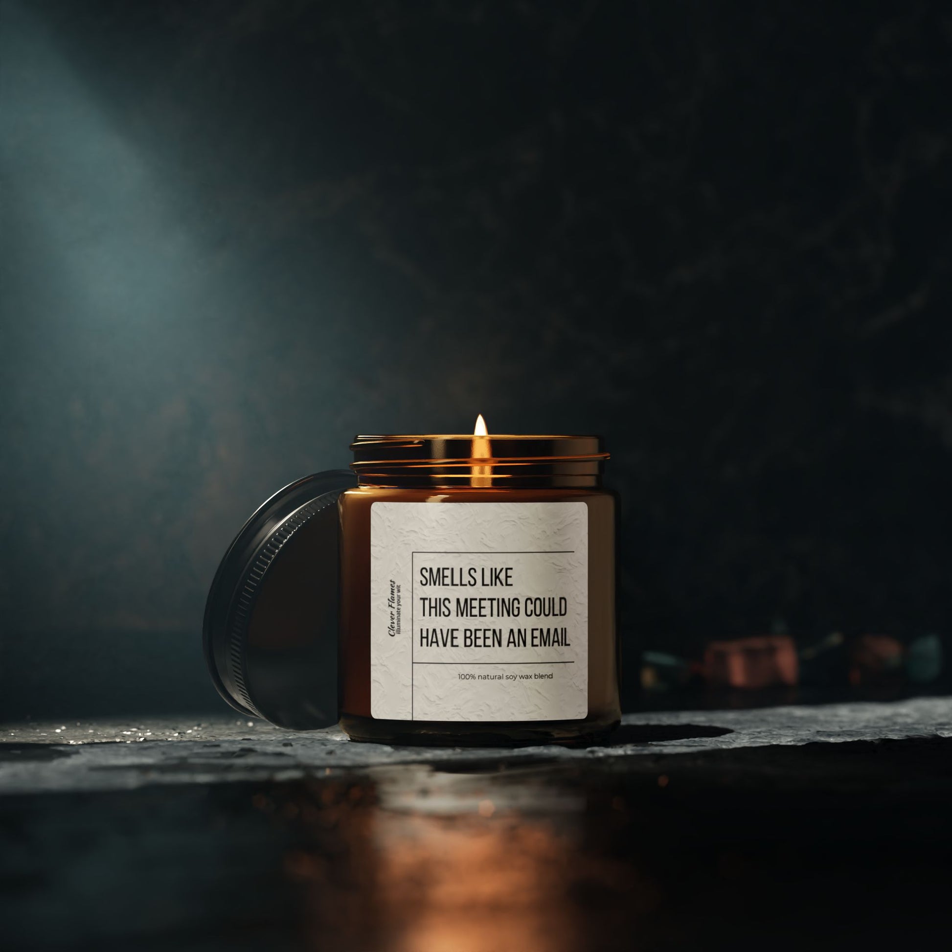 A glowing scented soy candle from Printify, designed to be eco-friendly, illuminates a dark setting. Its label humorously reads "Smells Like This Meeting Could Have Been an Email," making it the perfect gift for coworkers. The jar lid lies next to it on a textured surface.