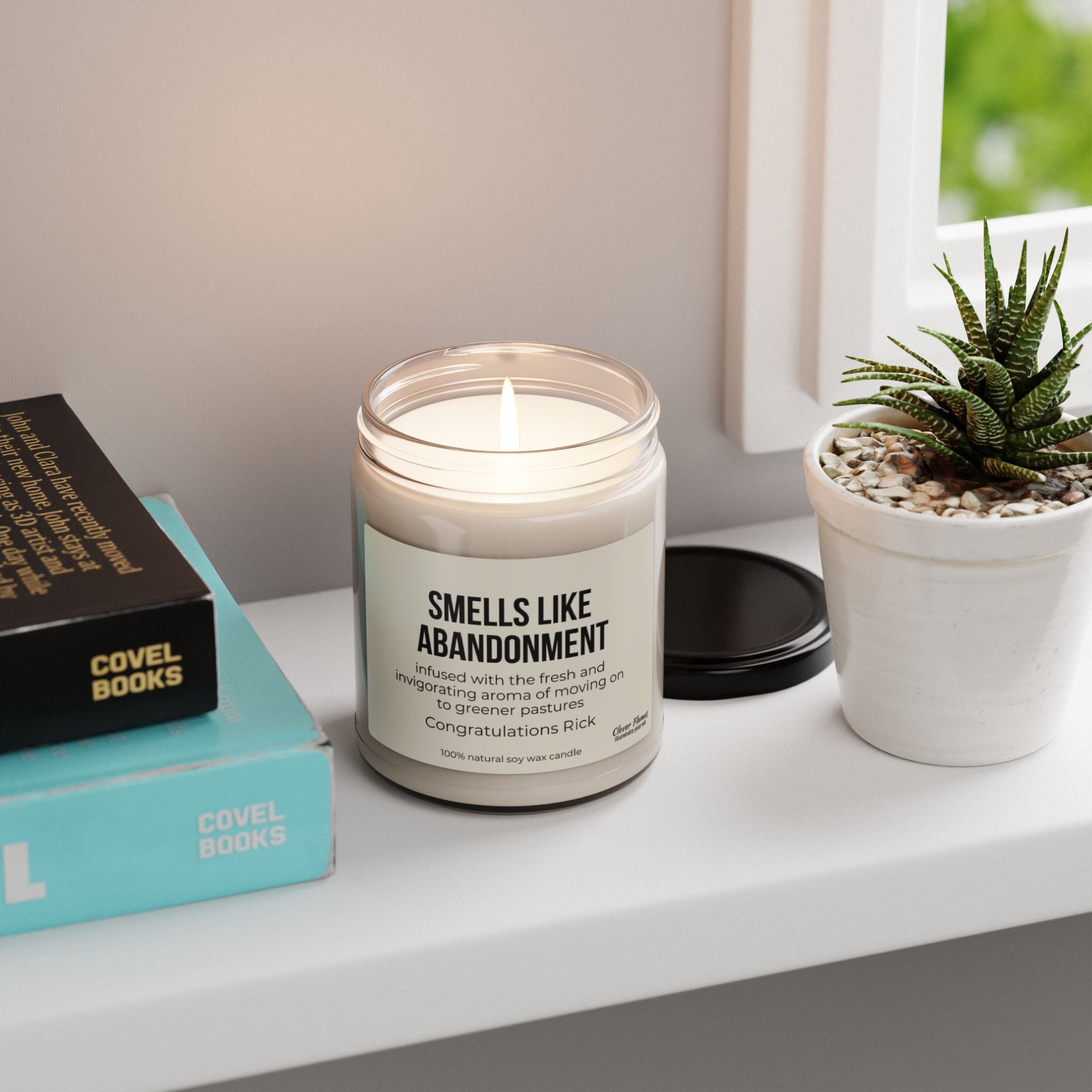 A lit soy candle from Printify, labeled "Abandonment Candle for Coworker Leaving Funny Gift Smells Like Retirement," sits on a white shelf next to a small potted succulent—an ideal gift for coworkers. Nearby are closed books with indistinct titles and a window revealing a blurred greenery view outside.