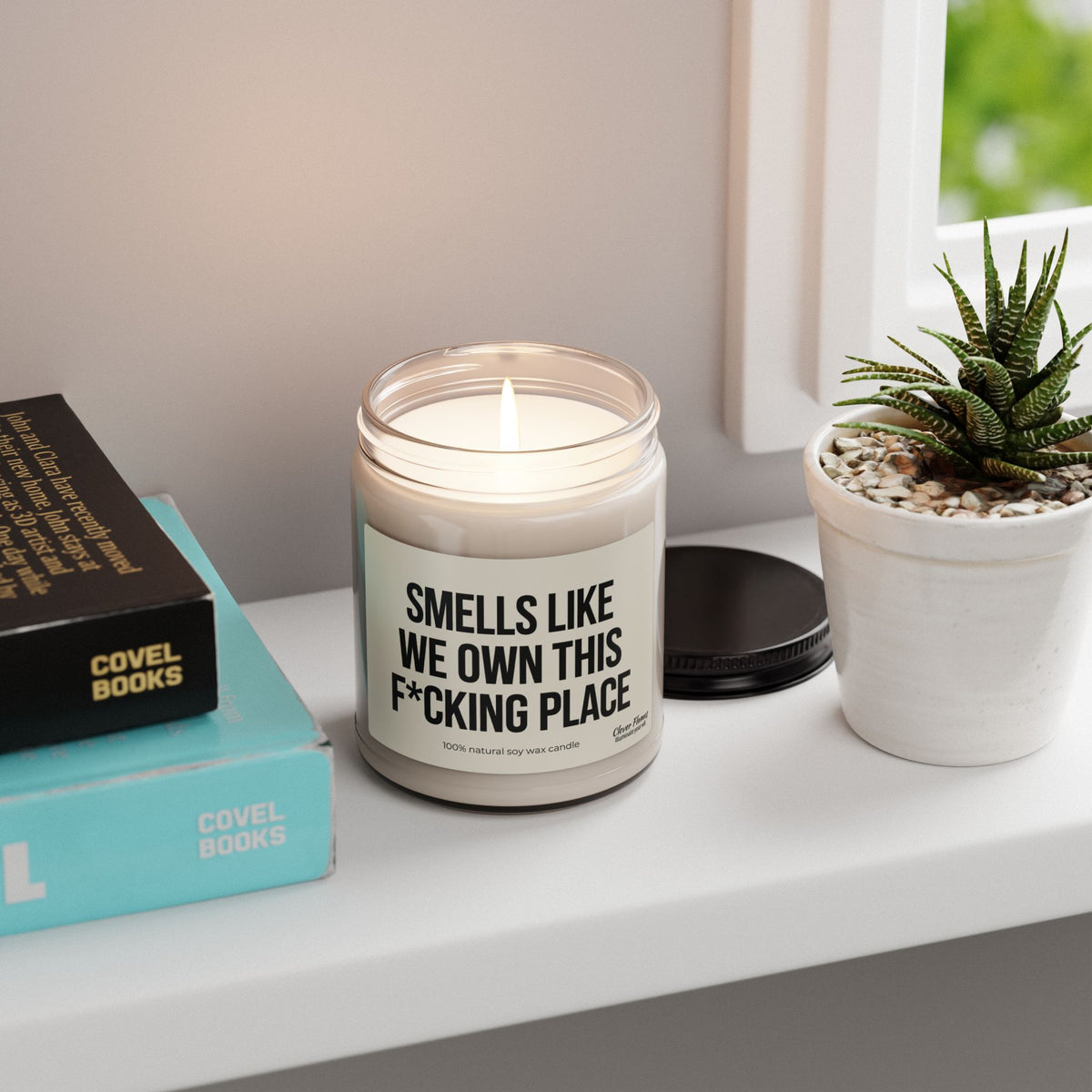 A lit Smells Like We Own This F*Cking Place Candle on a windowsill, with the label visible on the jar.