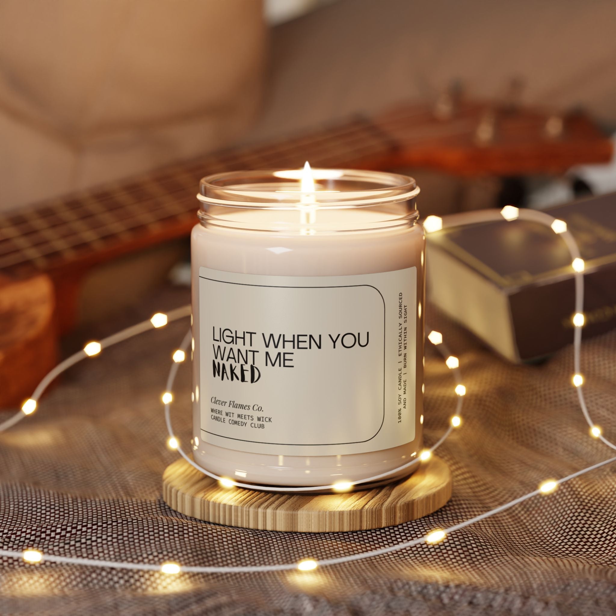 A lit candle in a glass jar, from the Printify collection, bears the label "Light When You Want Me Naked," and is crafted from natural soy wax, resting on a wooden coaster. It is surrounded by warm fairy lights, a plush pillow, a book, and part of a ukulele, creating an inviting and intimate ambiance—an ideal Valentine's Day gift for your boyfriend.