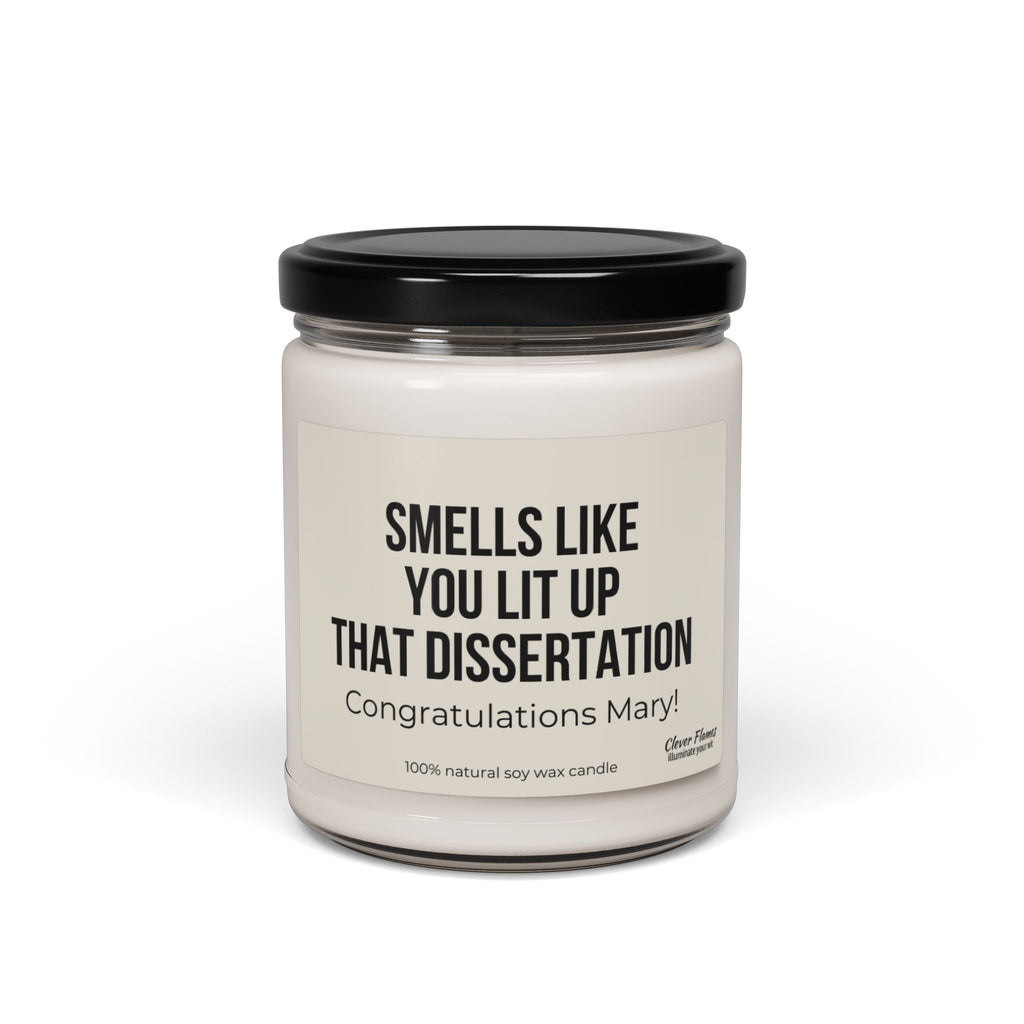 A personalized candle by Printify, featuring a white wax encased in a glass jar with a black lid. The label reads, "Smells like you lit up that dissertation. Congratulations Mary!" Made from 100% natural soy wax, this 'Smells Like You Lit Up That Dissertation' candle makes an ideal graduation gift for future doctors.