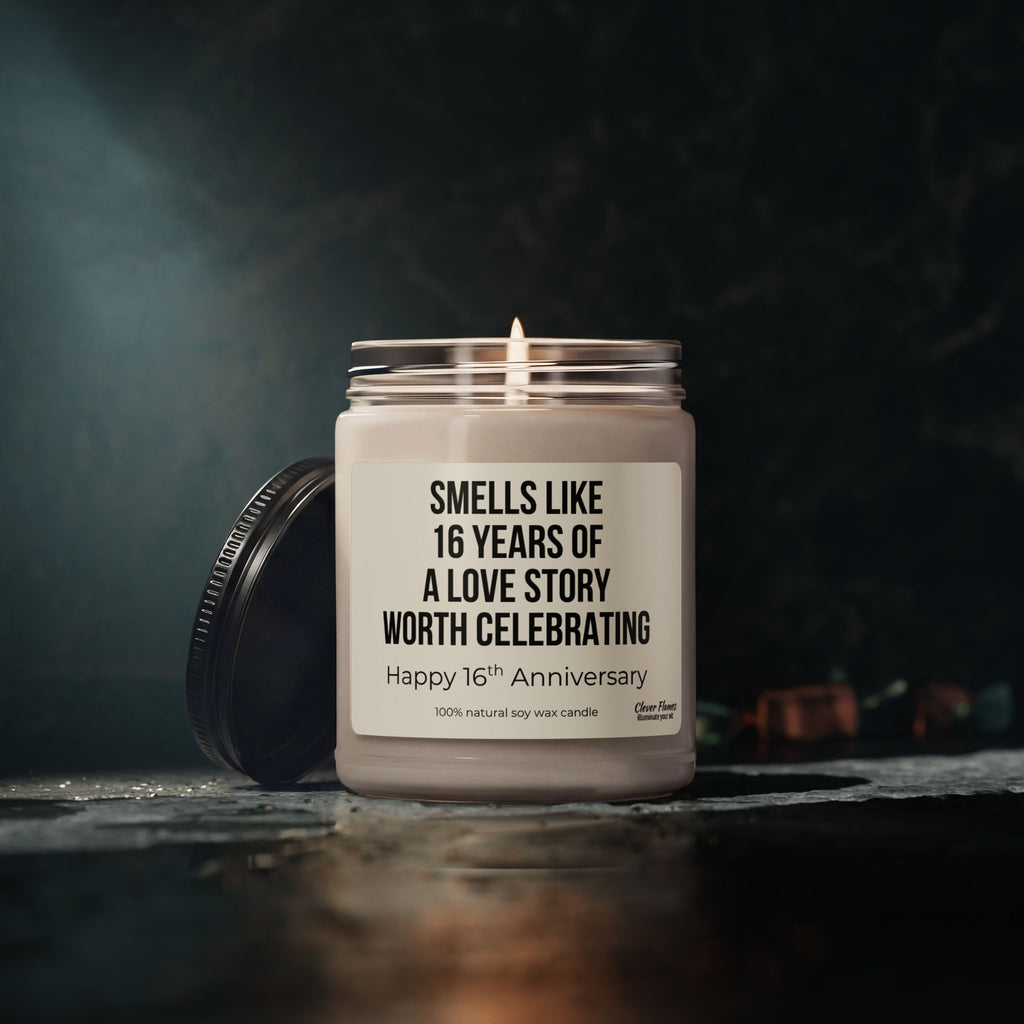 A lit Printify 16th Anniversary Candle sits in a glass jar, its label reading: “Smells like 16 years of a love story worth celebrating. Happy 16th Anniversary.” Crafted from 100% natural soy wax, this Wax Anniversary Gift for Parents or Couples is the perfect choice to commemorate 16 years married. The lid rests beside the jar.