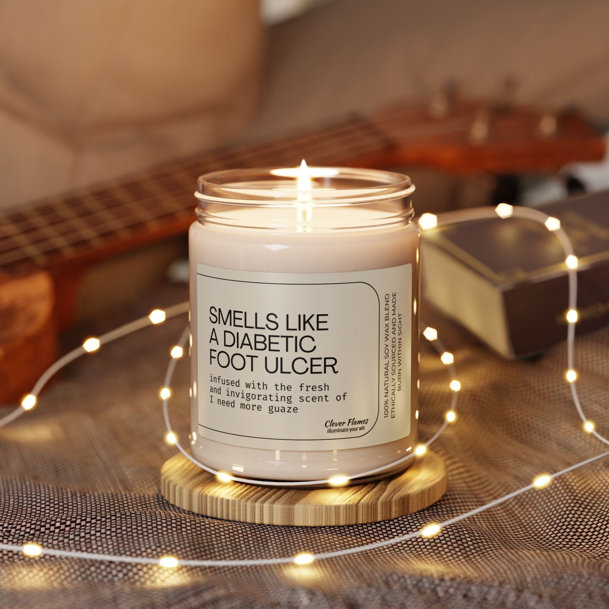The Funny Wound Care Nurse Candle by Printify, humorously labeled Smells Like a Diabetic Foot Ulcer, is lit on a wooden tray surrounded by fairy lights, part of a ukulele, and a book on cozy fabric—ideal for wound care nurses or those seeking funny medical gifts.