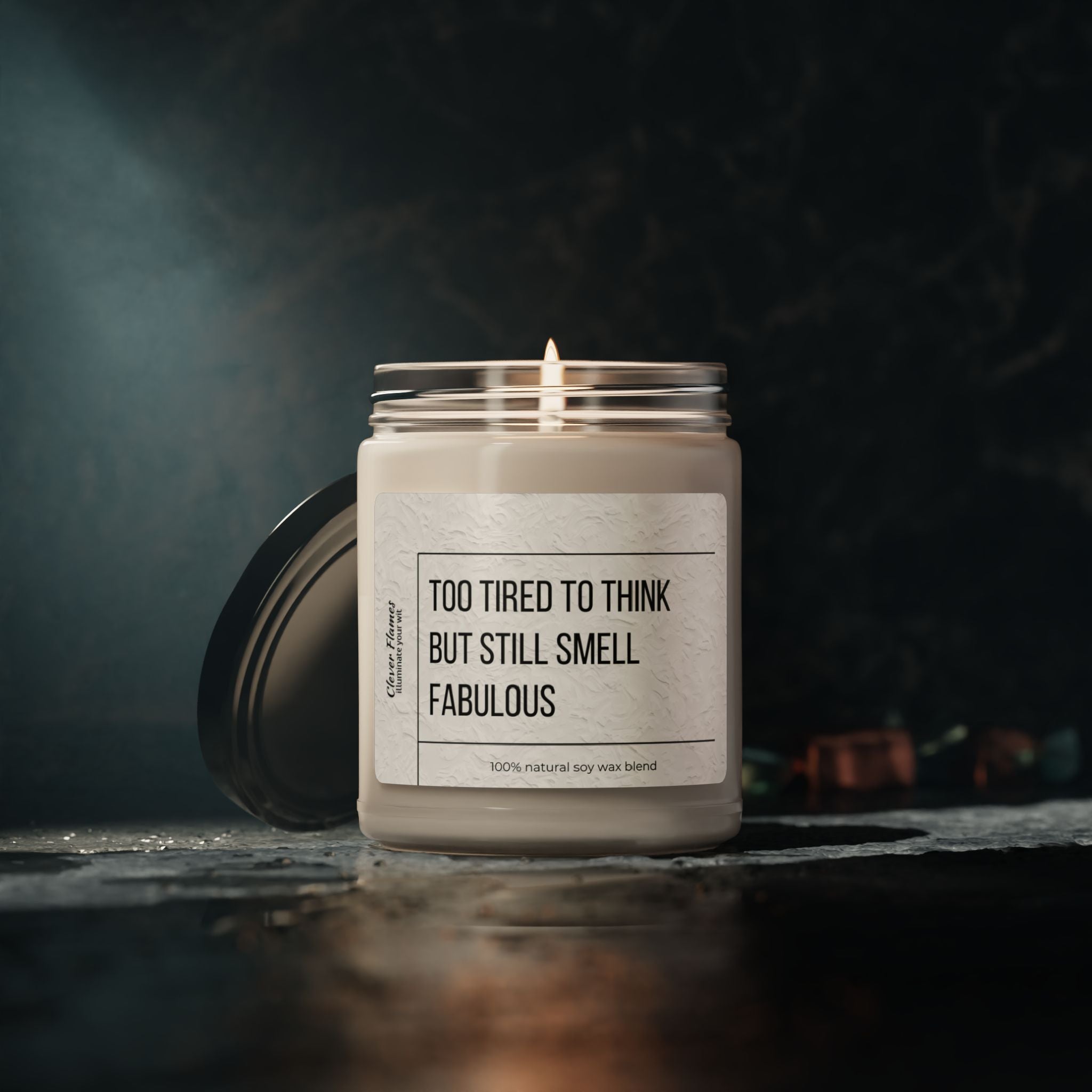 A lit soy candle from Printify, named "Too Tired to Think Candle - Smells Like Fabulousness," rests on a dark surface in a glass jar. Its label humorously declares its fabulous aroma even when you're too tired to think. This aromatherapy candle creates a soothing atmosphere with its gentle glow and relaxing, moody ambiance.