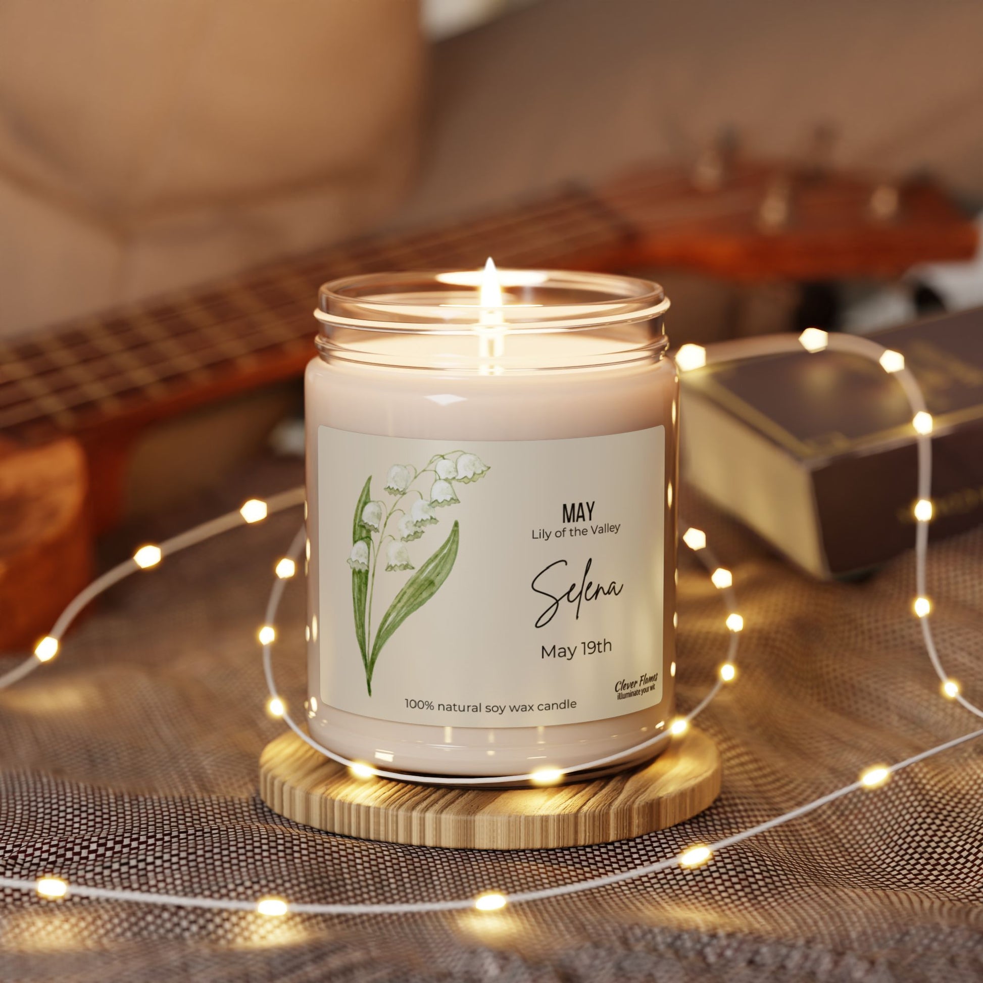 A personalized candle from Printify made of natural soy wax, sits in a glass jar with May, Lily of the Valley, Selena, May 19th and rests on a wooden coaster. Surrounded by fairy lights, its backdrop includes a blurred ukulele and book.