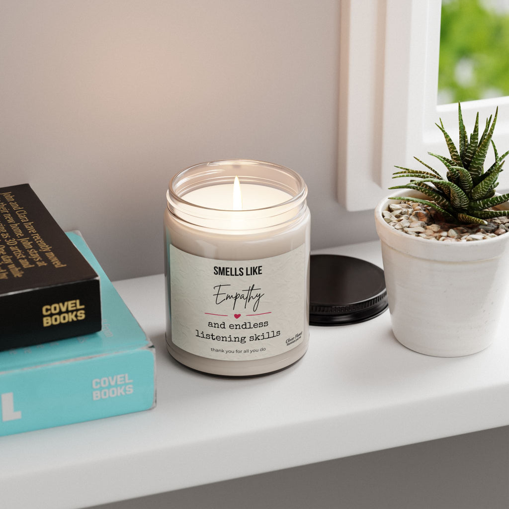 A 9oz Empathy Candle from Printify, labeled "Smells Like Empathy and Endless Listening Skills," sits on a white windowsill beside books, a black lid, and a small potted succulent as sunlight streams through the window.