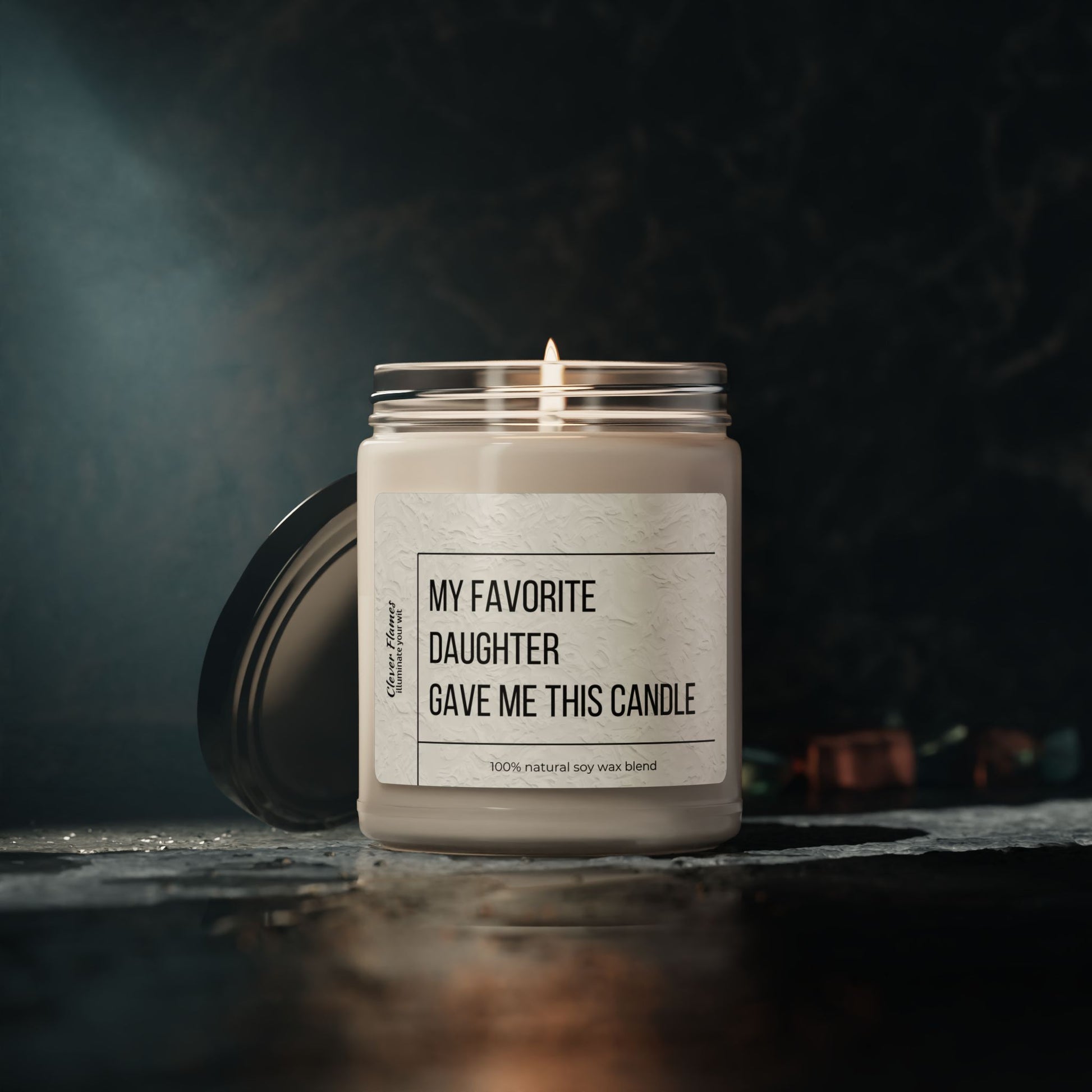 The Printify My Favorite Daughter Scented Soy Candle, 9oz, with a labeled glass jar and black lid, sits lit on a dark surface against a blurred background—ideal for birthdays, Mother’s Day, decor, relaxation, or celebrations.