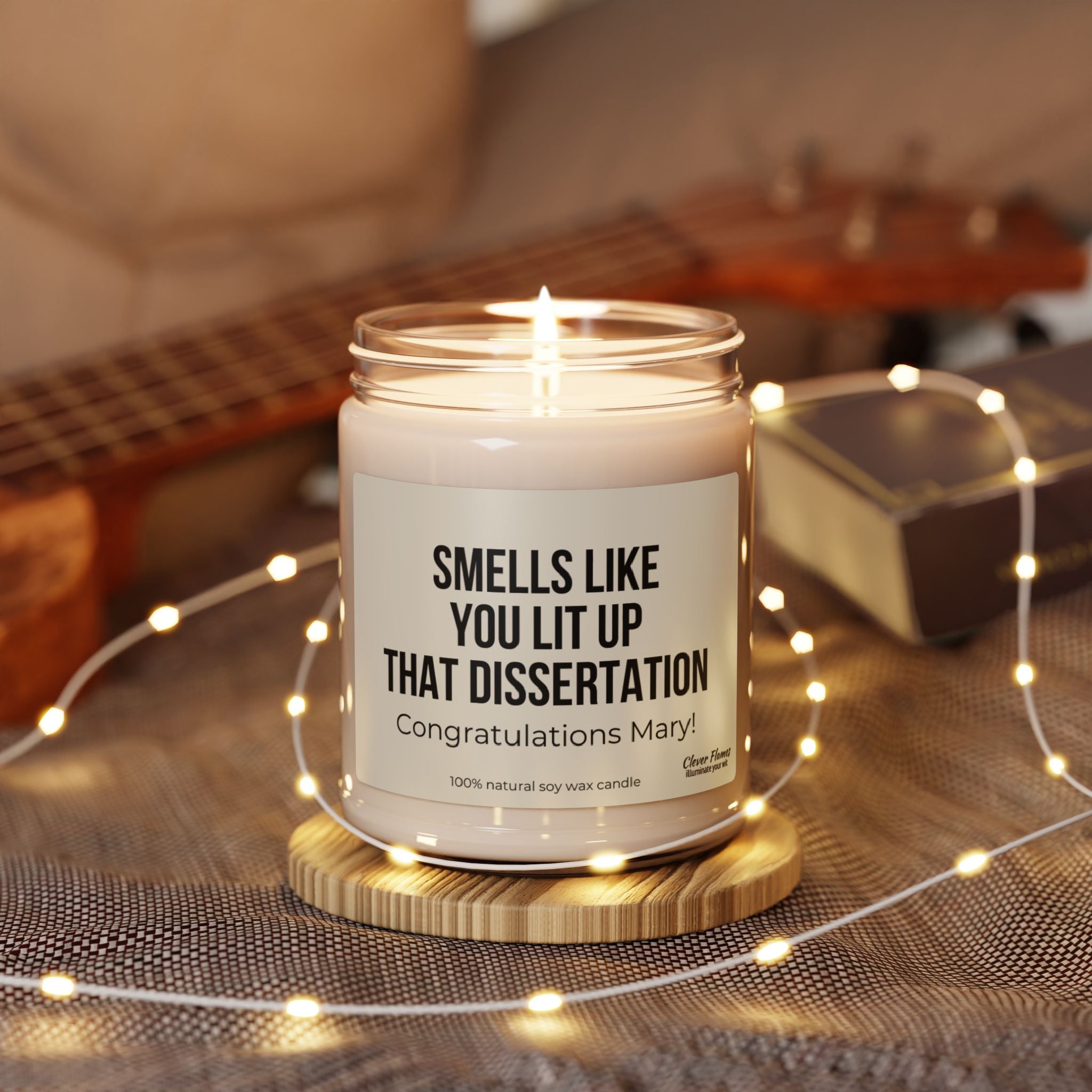 A personalized candle from Printify, labeled "Smells Like You Lit Up That Dissertation," rests on a wooden coaster, congratulating Mary on her doctorate achievement. Enveloped by fairy lights, the scene is complemented by a blurred ukulele and book in the background, making it an ideal gift for celebrating a future doctor.