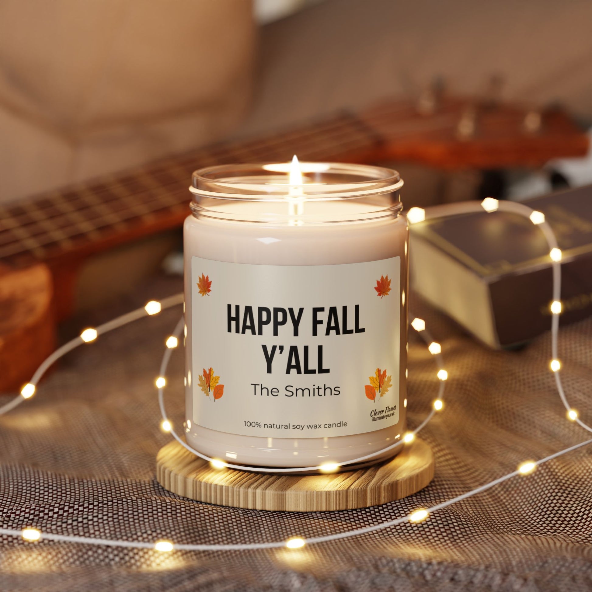 A personalized "Happy Fall Yall" scented soy candle from Printify, featuring the inscription "The Smiths," is placed on a wooden coaster surrounded by fairy lights. In the background, a woven fabric and a ukulele case add to the cozy autumn atmosphere, making for ideal fall decor.
