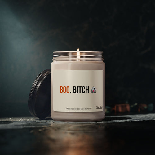 A lit Printify Funny Halloween Fall Candle sits in a glass jar labeled “BOO. BITCH” with a ghost emoji. Its black lid rests nearby on a dark, moody backdrop, making it the ultimate spooky and sarcastic autumn decor or party favor—perfect for gifting sass and atmosphere to her or your bestie this fall season.