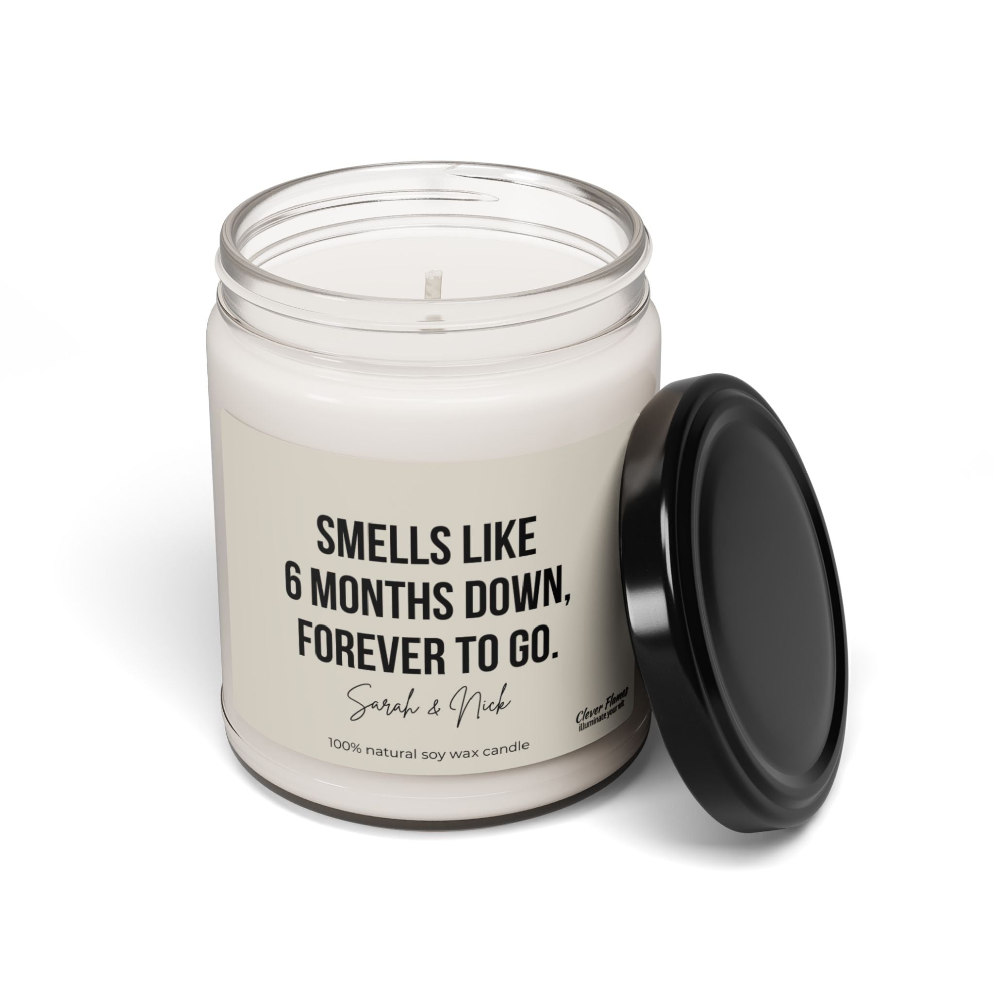 A white candle in a glass jar with a black lid beside it commemorates a 6 months anniversary. The label reads, "Smells like 6 months down, forever to go. Sarah & Nick," adding a personalized touch. It notes it is made from 100% natural soy wax at the bottom. This candle, by Wedding, is the perfect personalized 6 Month Anniversary Gift and can also be given as a funny gift for friends, spouses, or best friends on their birthday or Valentine's Day.