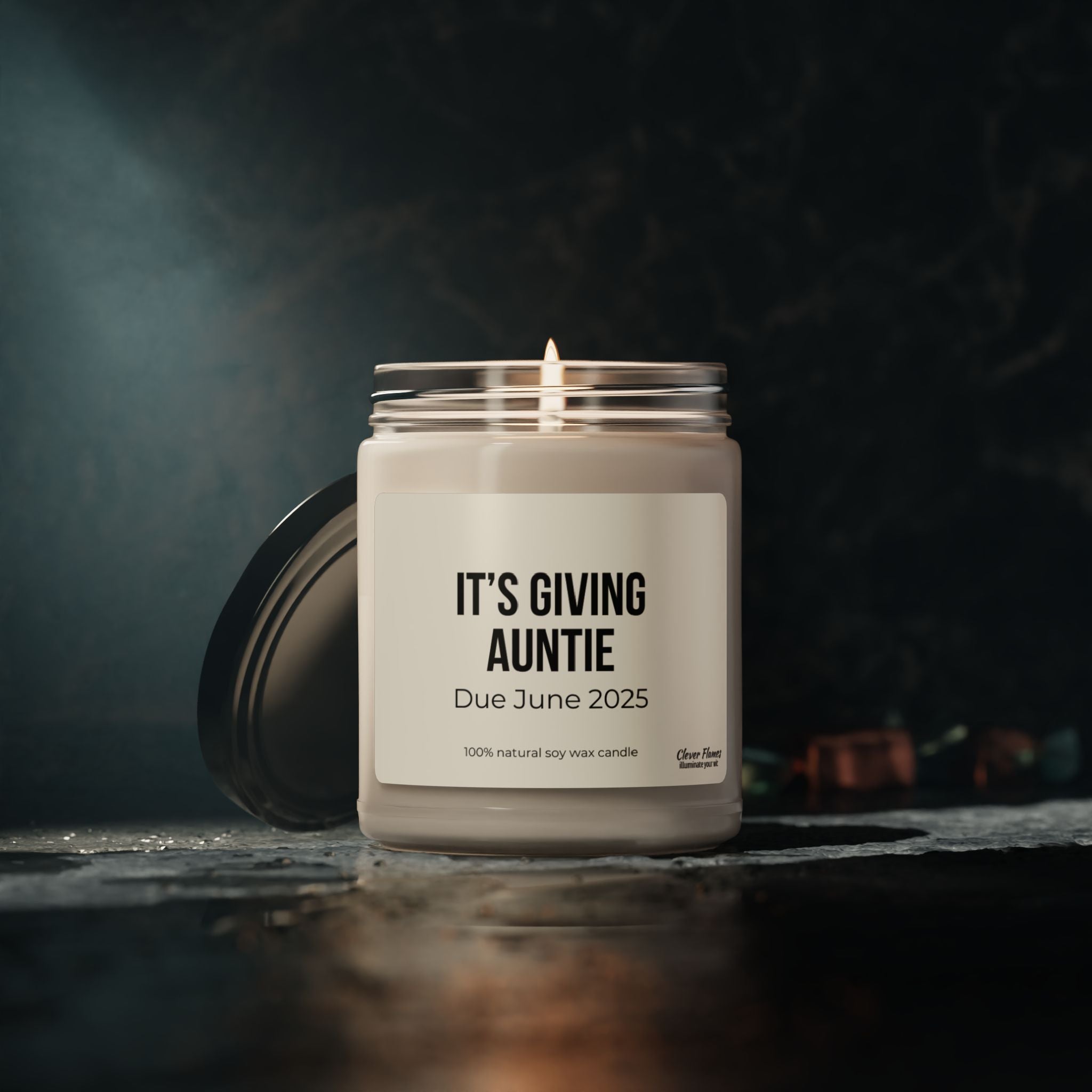 A lit soy wax candle in a jar from Printify features the label ITS GIVING AUNTIE, with a due month of June 2025. This customizable scented candle is perfect for baby reveal celebrations, offering a dark and moody ambiance.