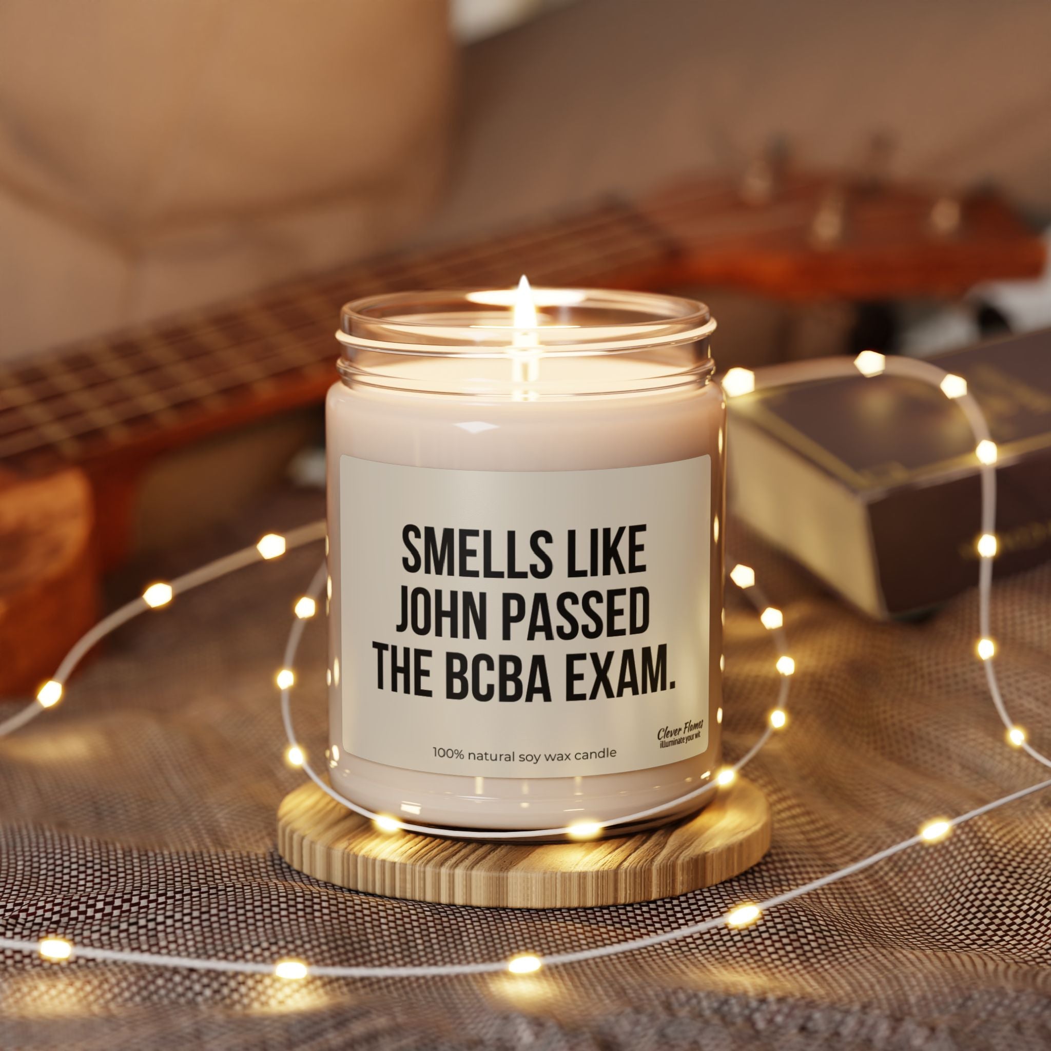 A Personalized Behavior Analyst Candle Gift from Printify, designed for celebrating passing the BCBA exam, rests elegantly in a jar atop a wooden coaster. Its label humorously states "Smells like John passed the BCBA exam." Made from a natural soy wax blend, this candle enhances the cozy ambiance of a background adorned with a guitar and string lights.