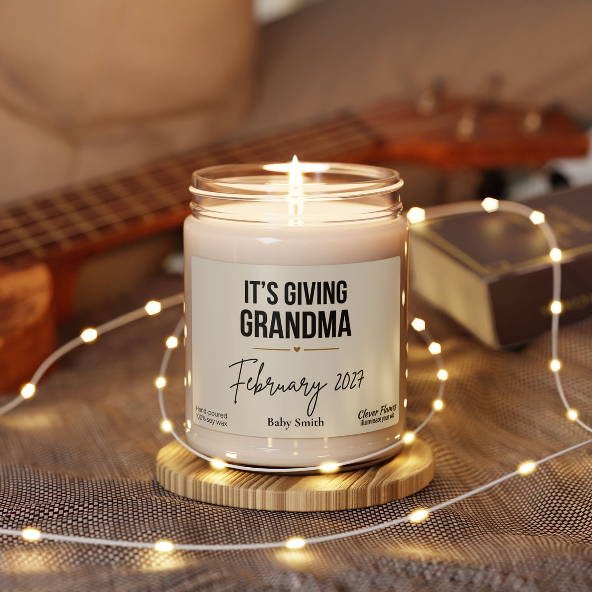 A lit Grandma Pregnancy Announcement Candle in a glass jar, featuring a label that reads Its Giving Grandma, February 2027, Baby Smith, sitting on a wooden coaster surrounded by string lights.
